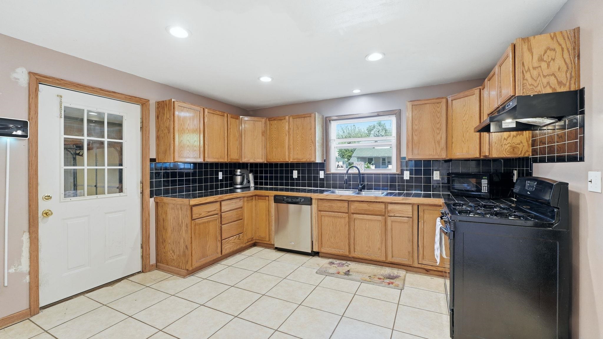 Kitchen featuring wood-finish cabinetry, black tile backsplash, stainless steel dishwasher, and a four-burner range with overhead microwave