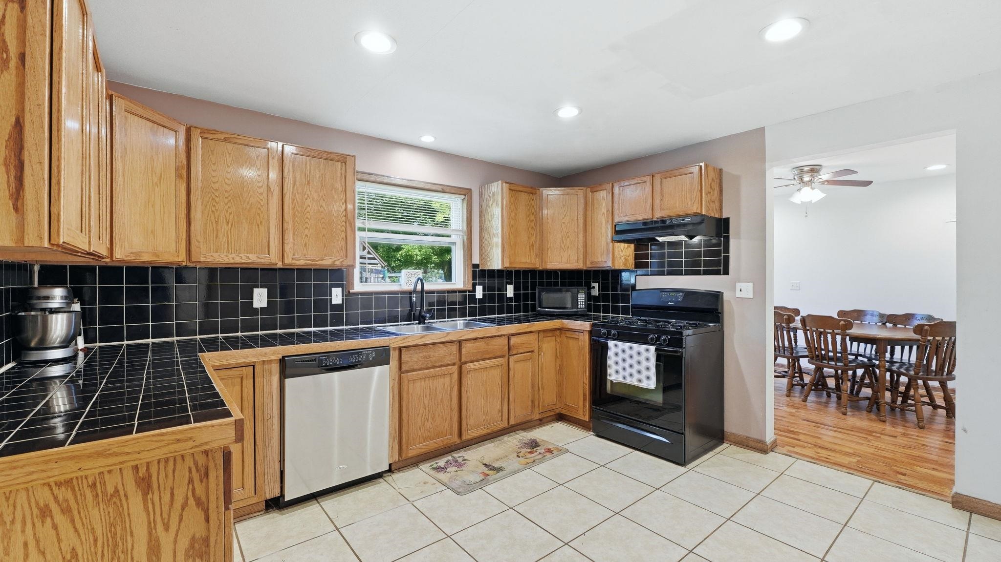 Kitchen featuring light wood cabinetry, black tile countertops, black tile backsplash, stainless steel dishwasher, and a black gas range