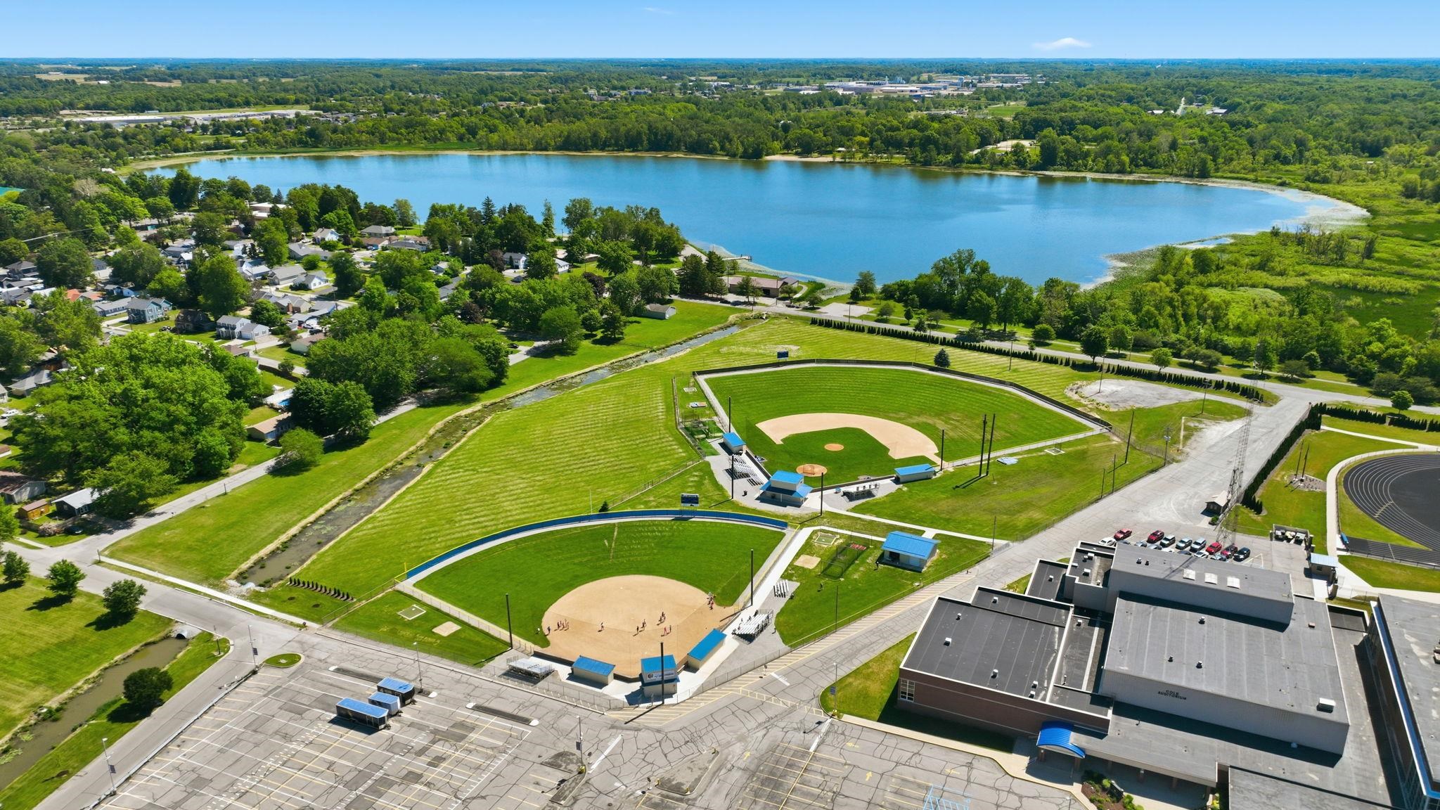 Expansive aerial perspective featuring a large freshwater lake, two baseball fields, and a sports complex building