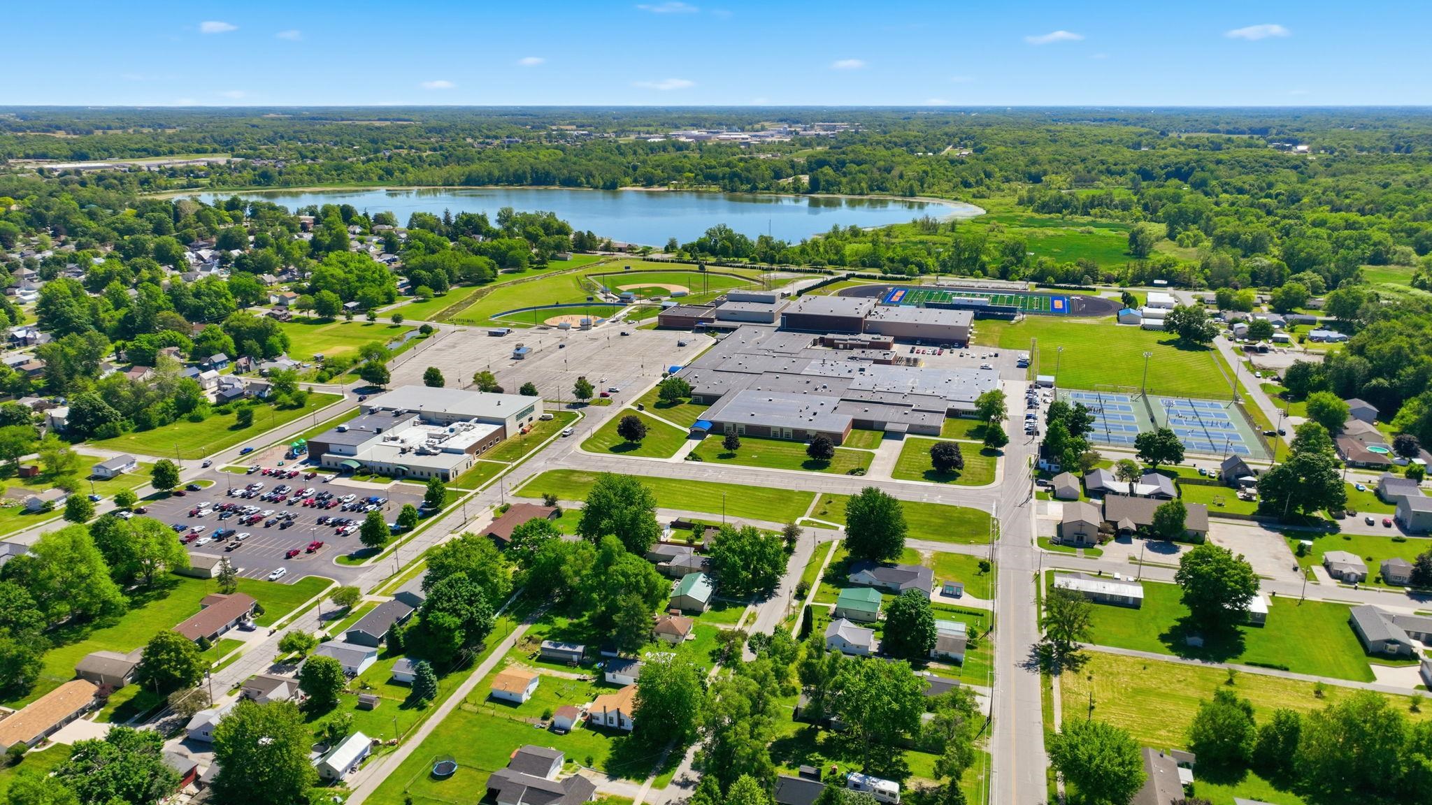 Expansive aerial perspective showcasing a residential area with a prominent body of water, extensive green spaces, and a large institutional complex featuring multiple buildings and athletic courts