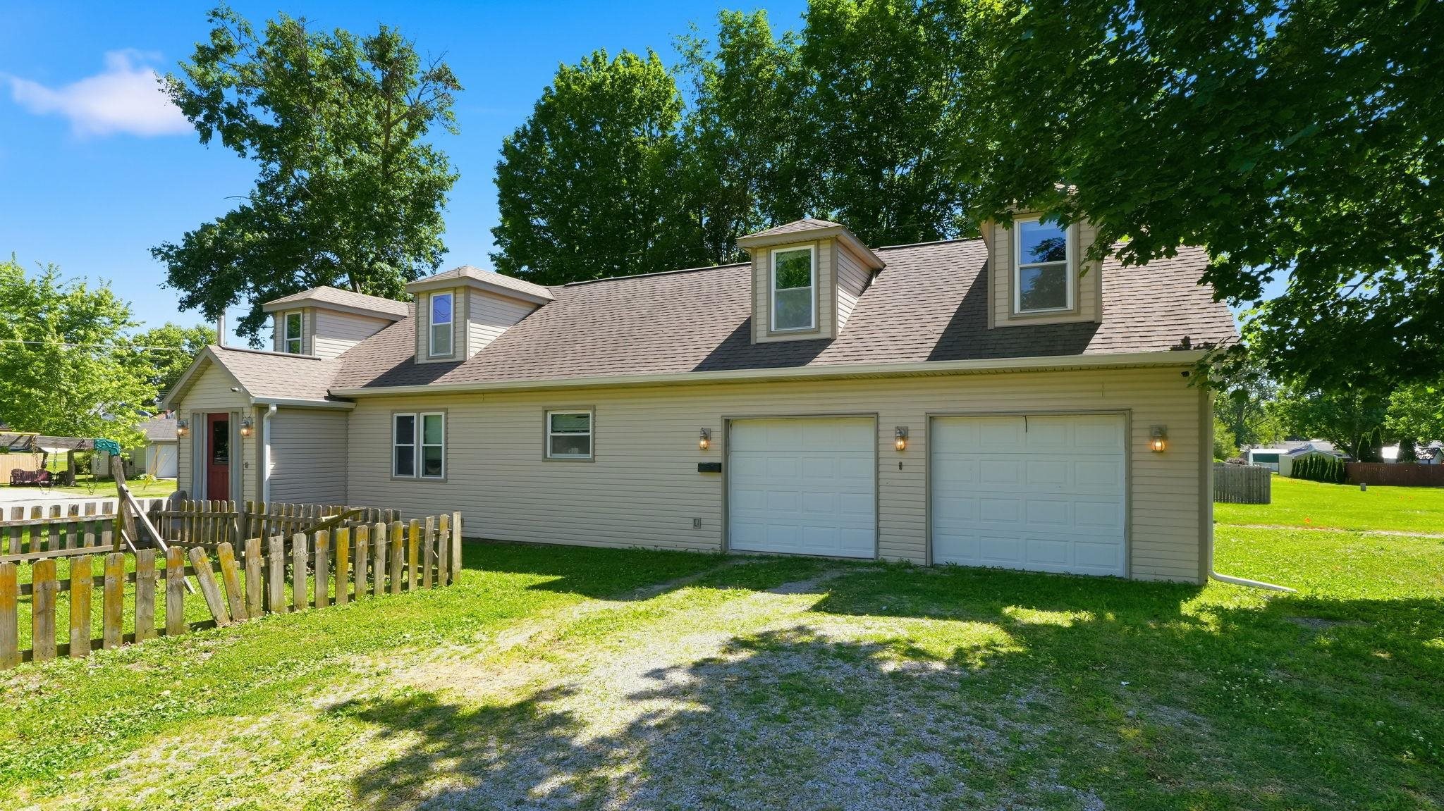 Two-car garage with overhead lighting, multiple dormer windows, a red entry door, and a partially fenced front yard
