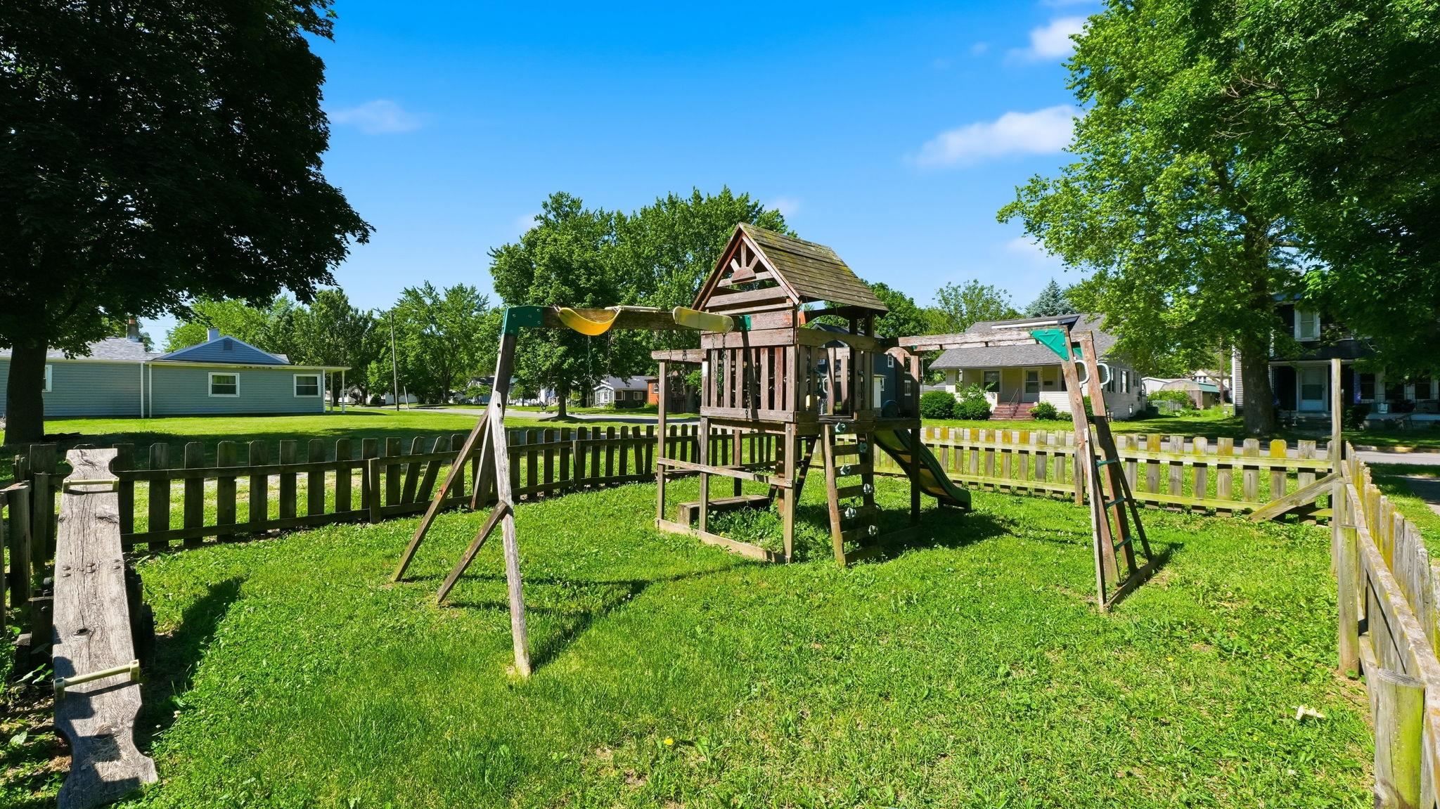 Expansive lawn area featuring a wooden play structure with a slide and swings, surrounded by a wood picket fence