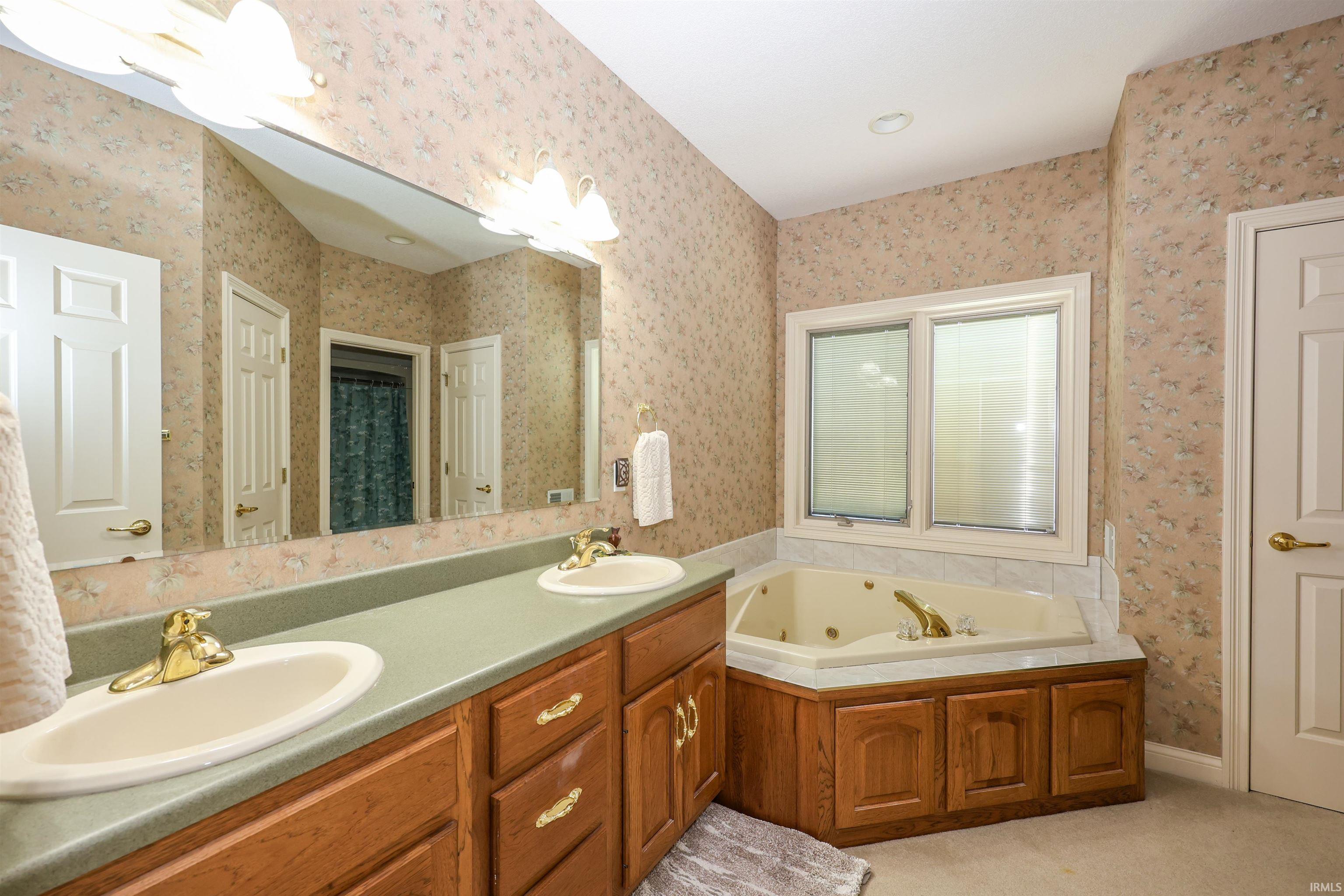 Bathroom featuring a double vanity with wood cabinetry, a large soaking tub with a tiled surround, an oversized mirror, and overhead lighting