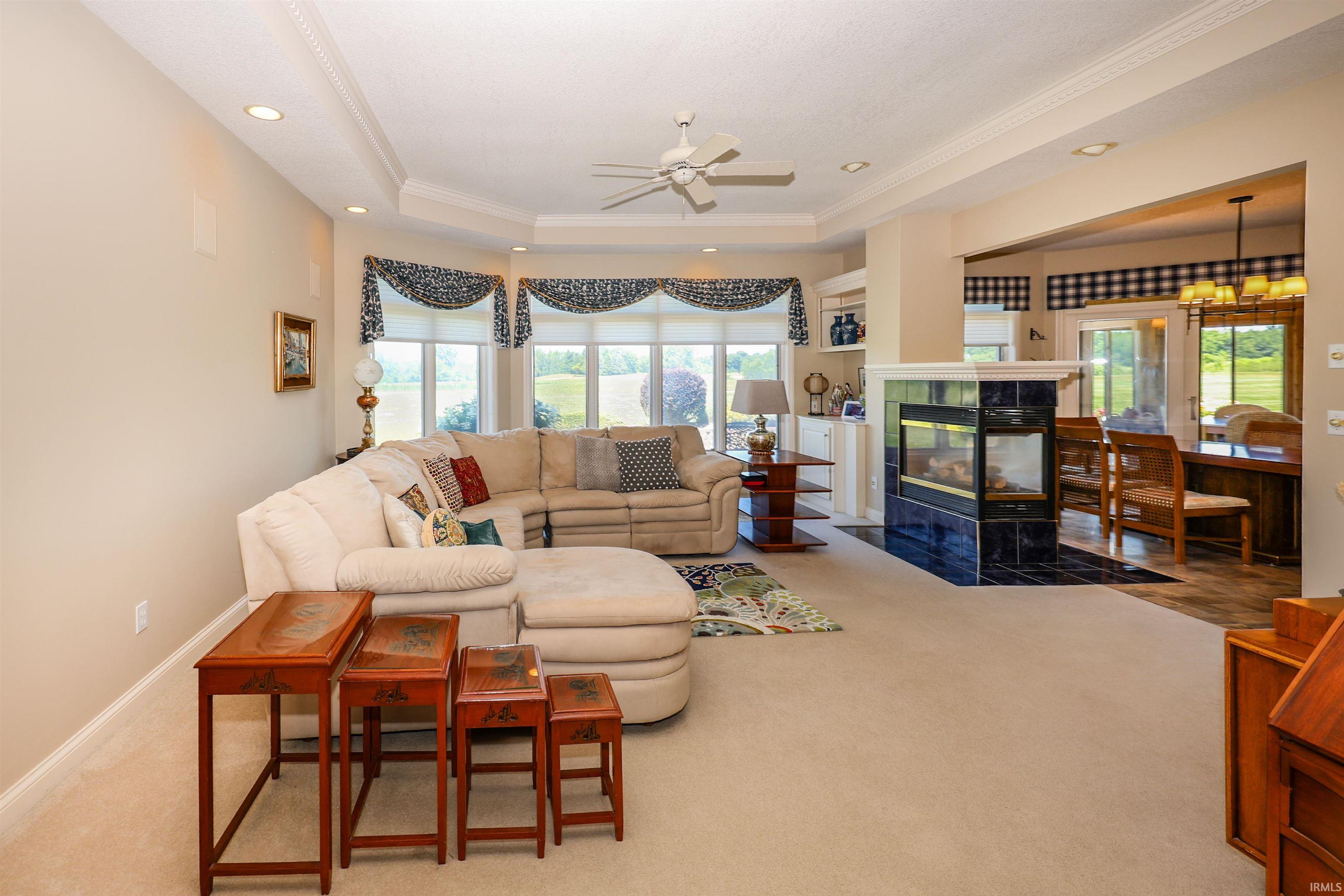 Spacious living area featuring a two-sided fireplace with dark tile surround, tray ceiling, recessed lighting, and large windows