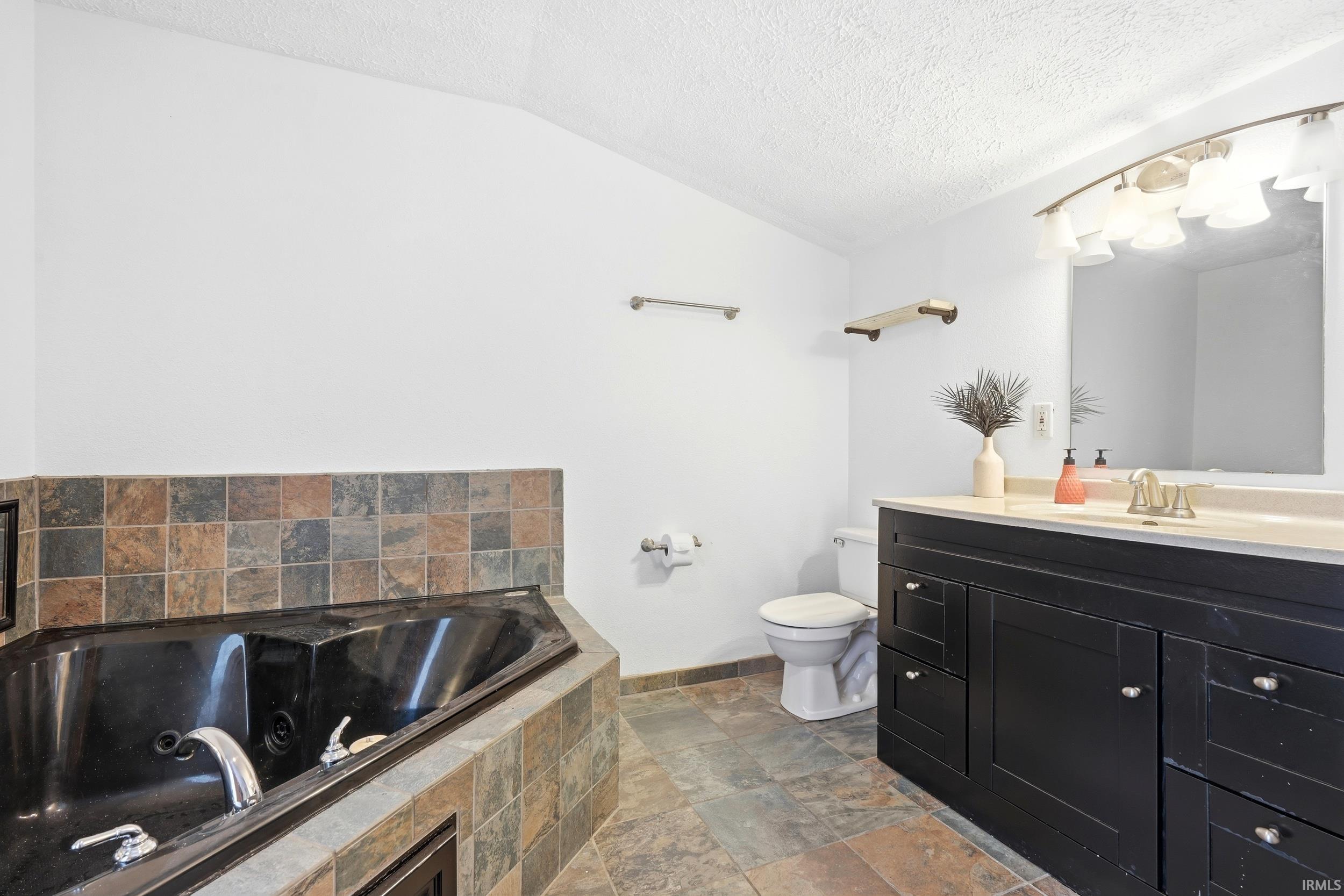 Spacious bathroom featuring a corner jetted tub with tile surround, a dark wood vanity with a light countertop, a toilet, and coordinating tile flooring