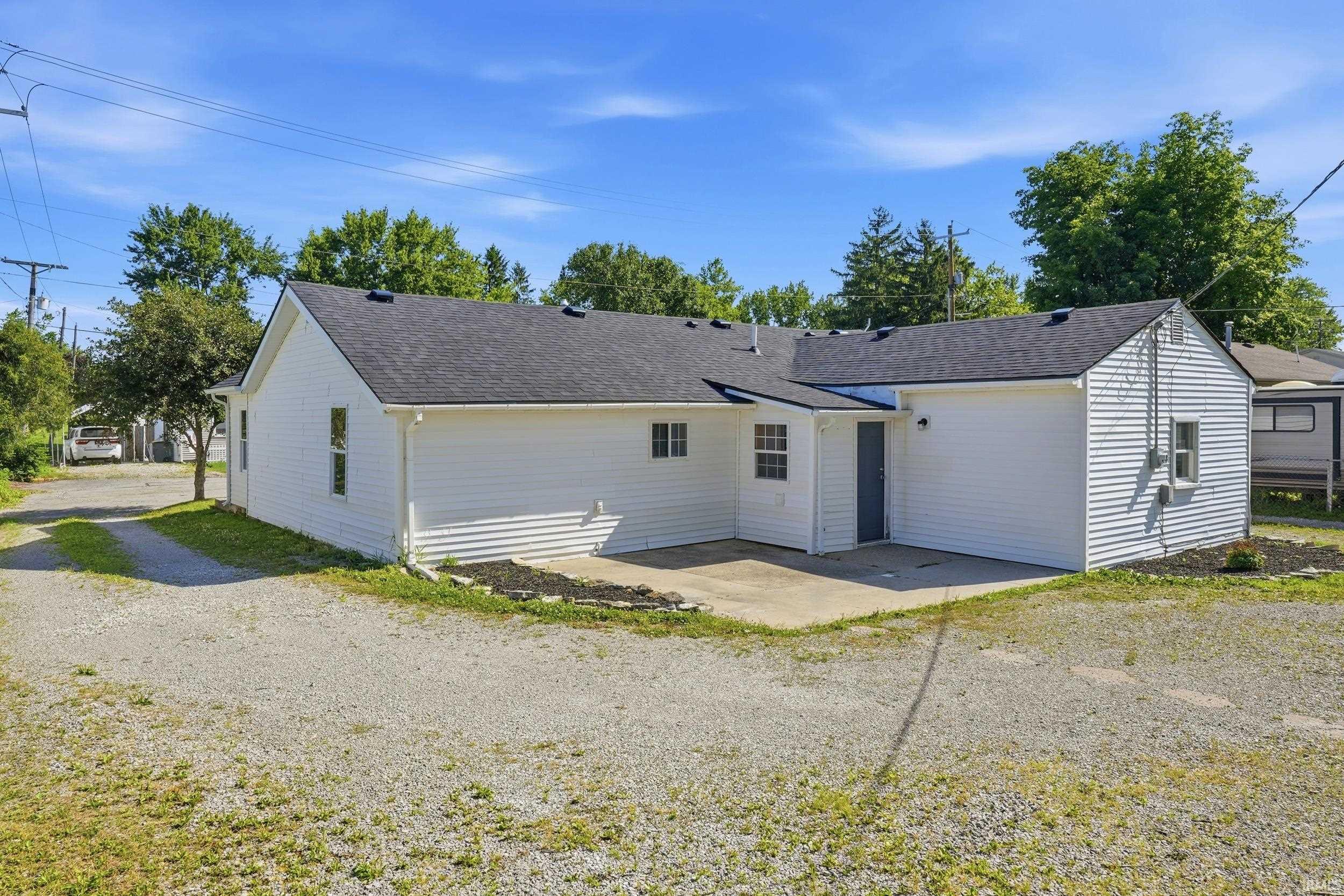 Rear exterior featuring white horizontal siding, a dark shingle roof, a concrete patio, and a gravel yard