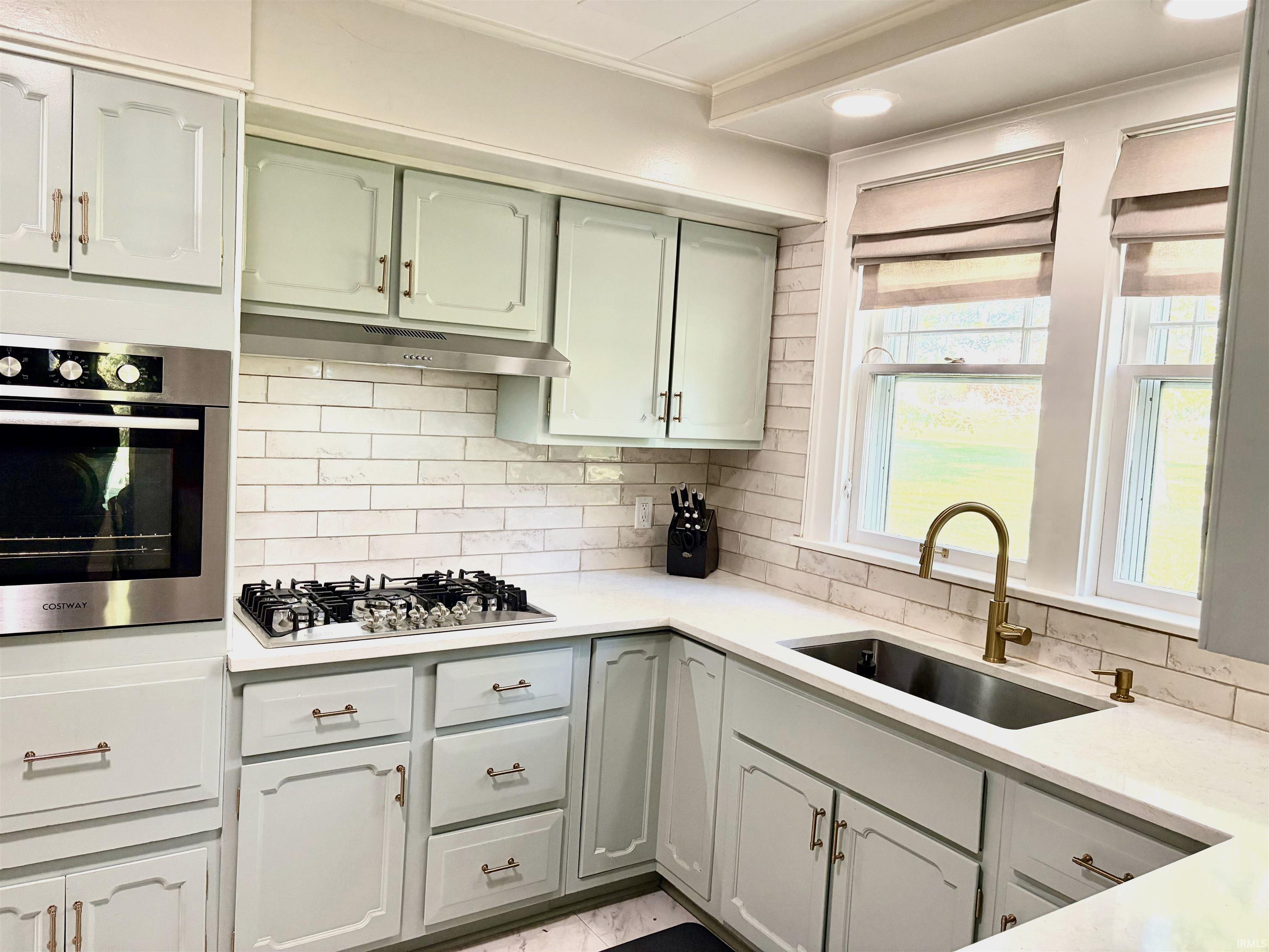 Kitchen featuring light-toned cabinetry with brass-toned hardware, white solid surface countertops, and a subway tile backsplash