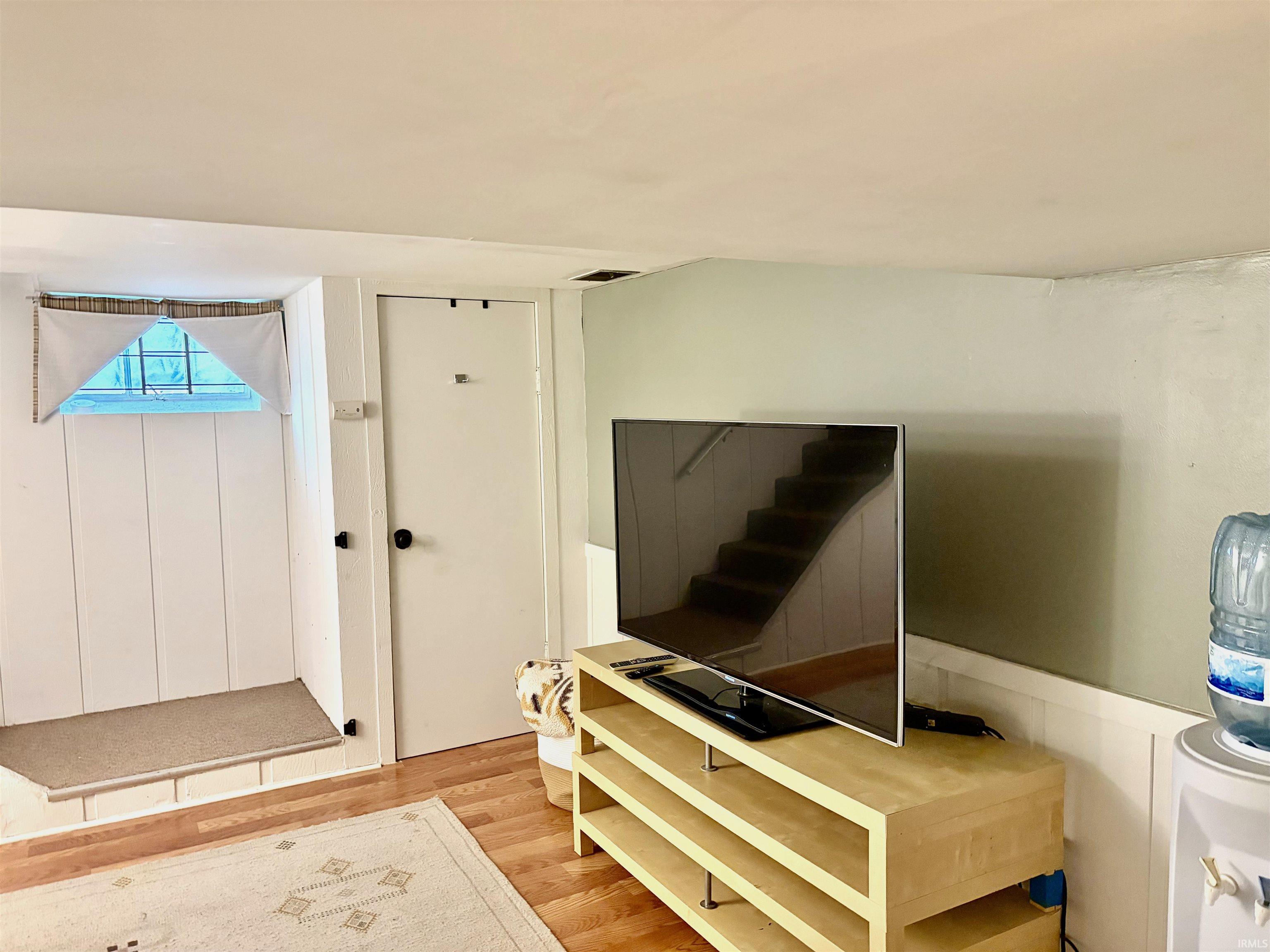 Basement area featuring a window with natural light, a white paneled wall, a white door, and wood-finish flooring