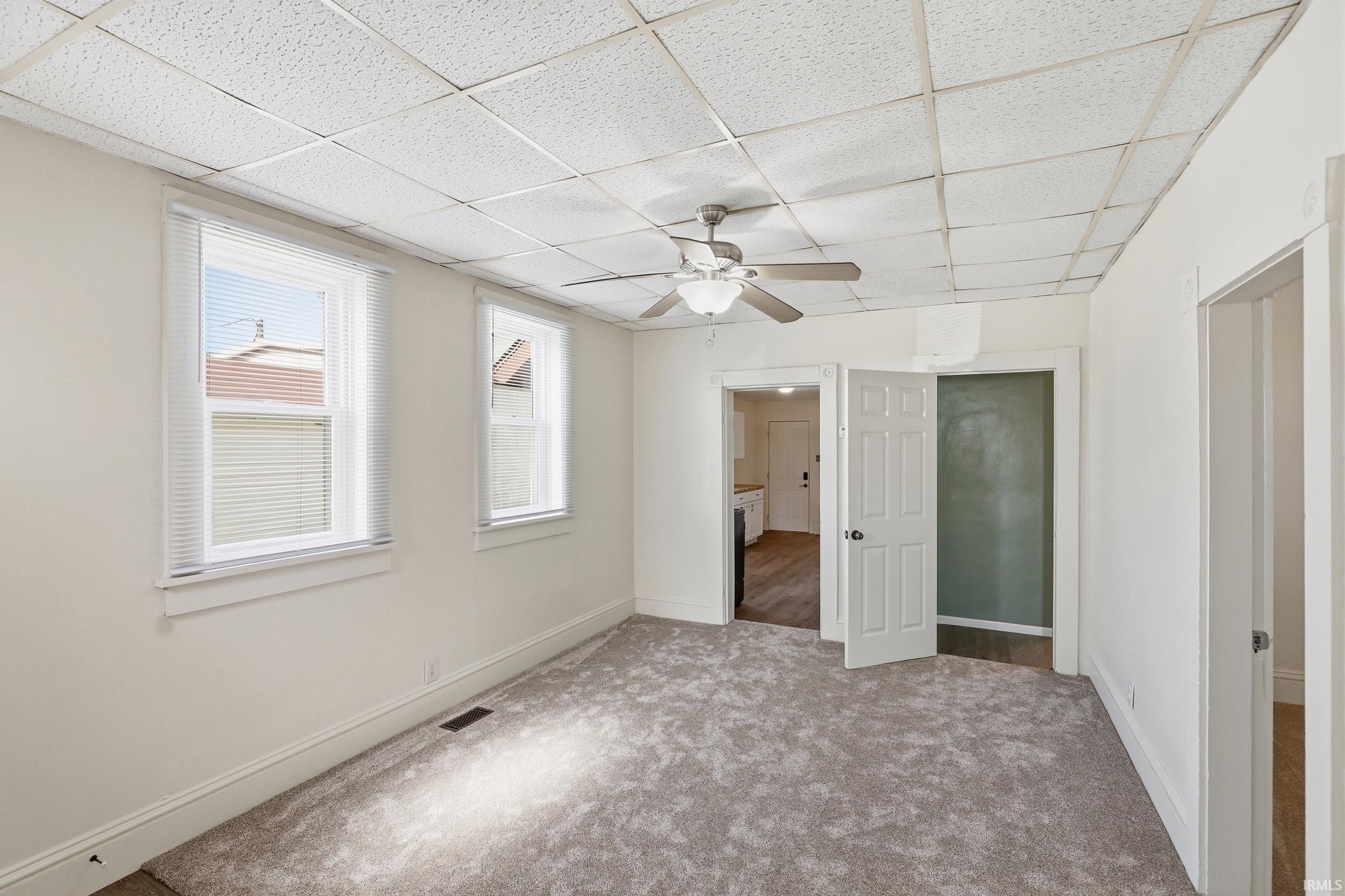 Carpeted room featuring a ceiling fan, two windows with blinds, white trim, and a drop ceiling