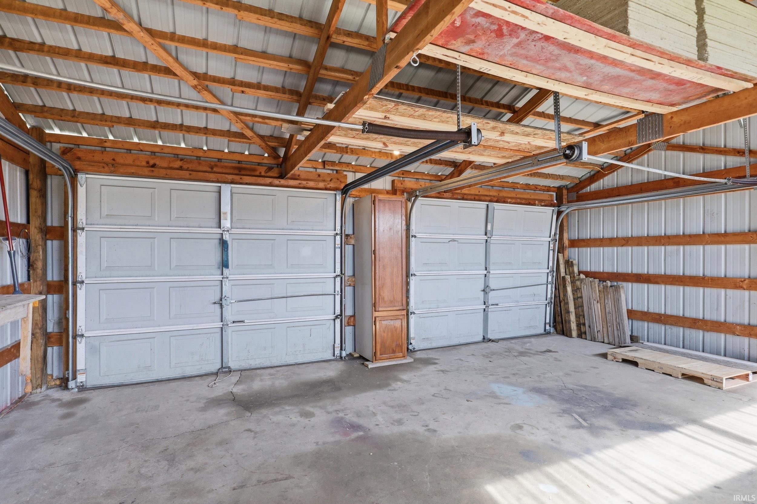 Two paneled garage doors set within a structure featuring a concrete floor, exposed wood beam ceiling, and metal wall siding