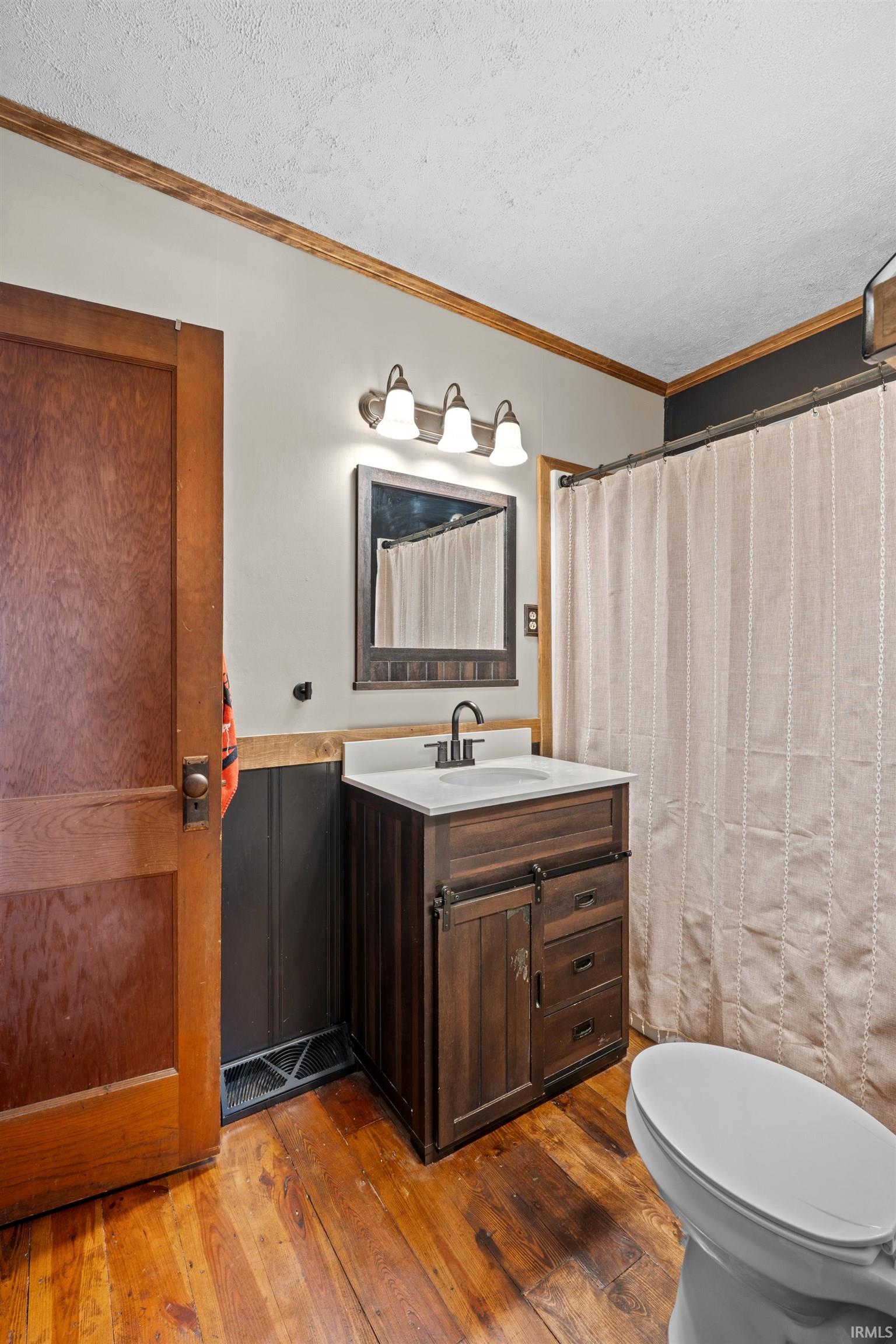 Bathroom featuring wood-finish flooring, a dark wood-tone vanity with a white countertop and integrated sink, and a framed mirror illuminated by a three-bulb light fixture