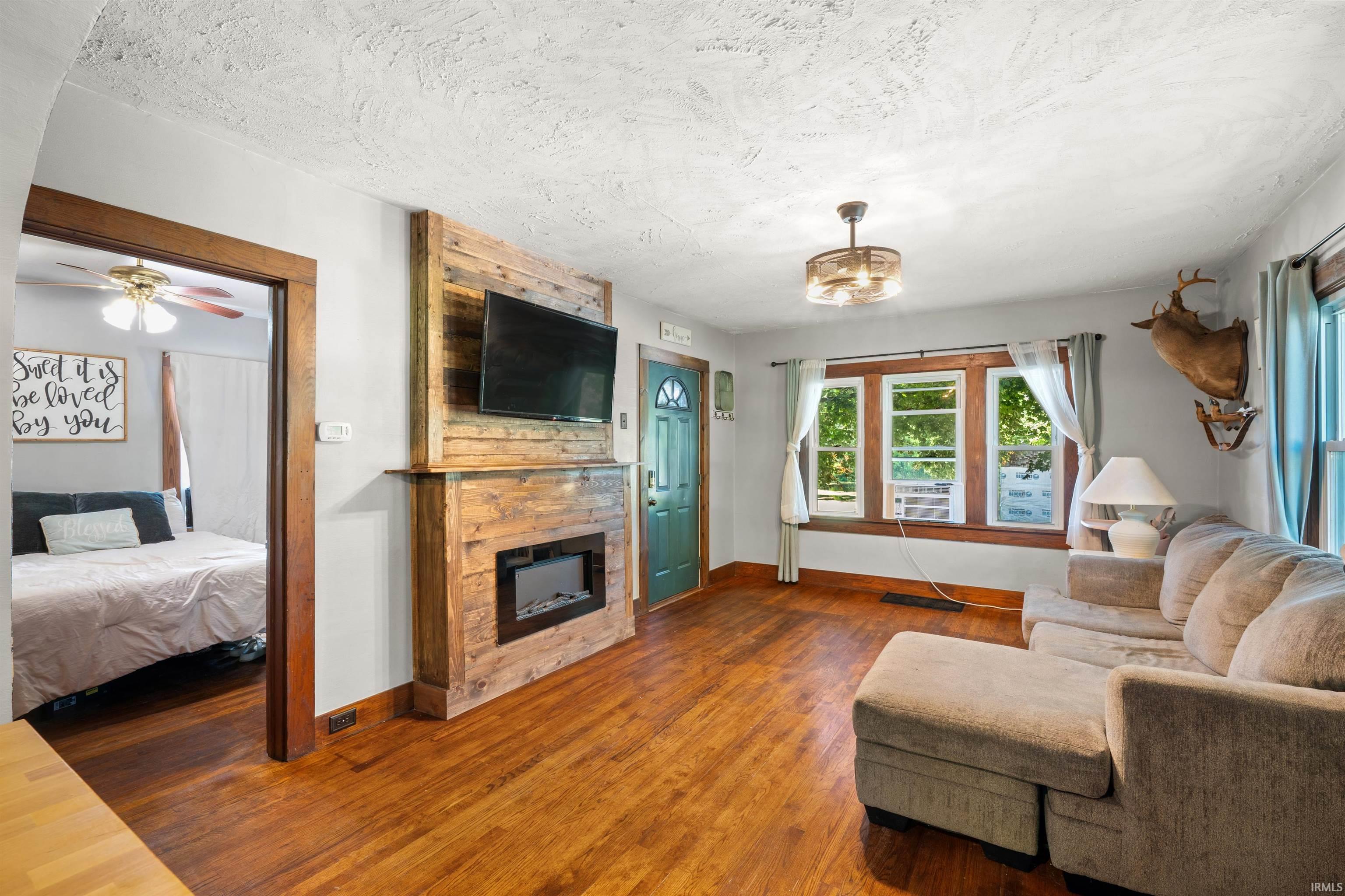Living area featuring wood-finish flooring, a wood-paneled accent wall with an electric fireplace, a ceiling fan, and a turquoise entry door with an arched window