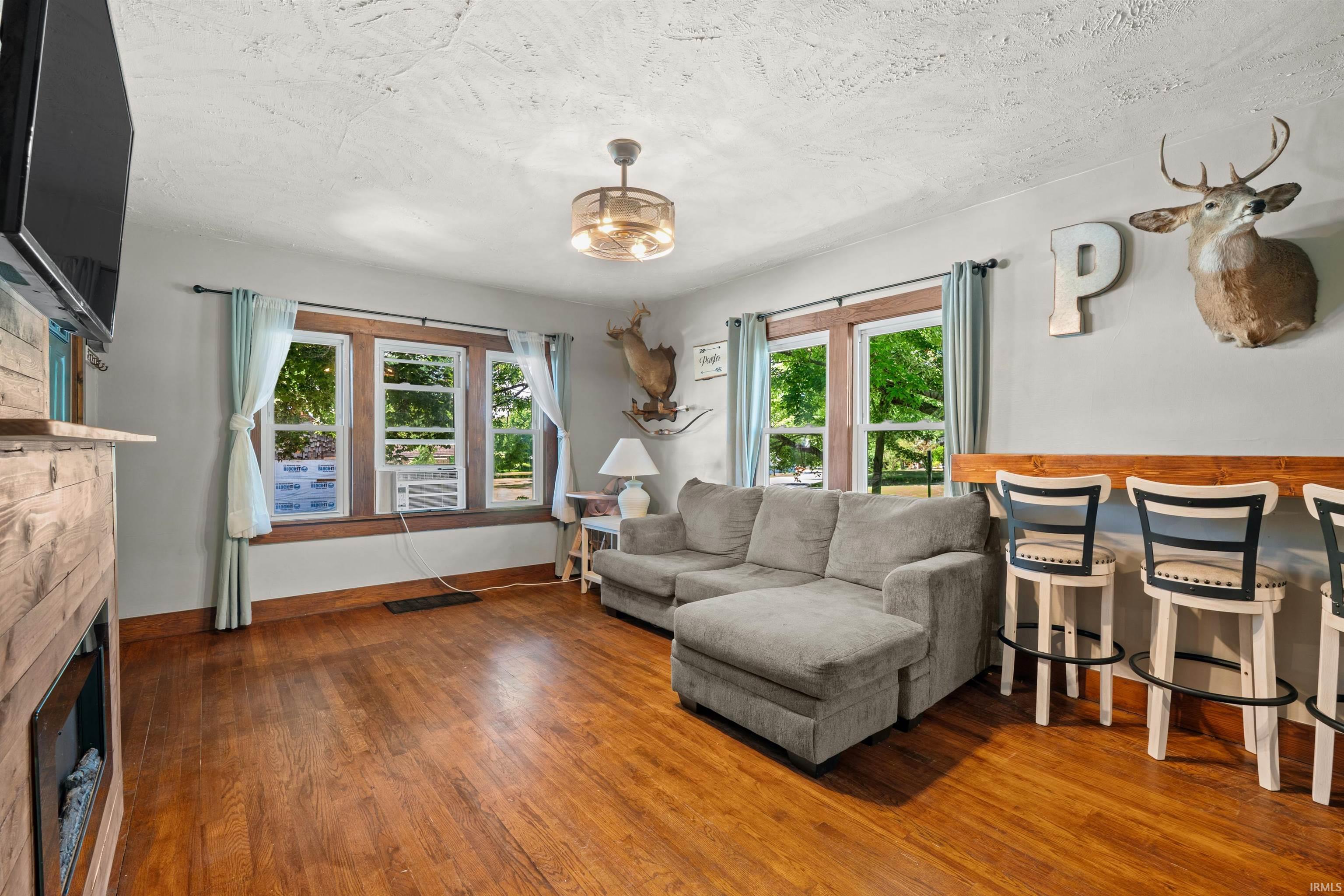 Spacious living area featuring wood-finish flooring, a built-in media console with an electric fireplace, multiple windows with wood trim, and a ceiling fan with integrated lighting