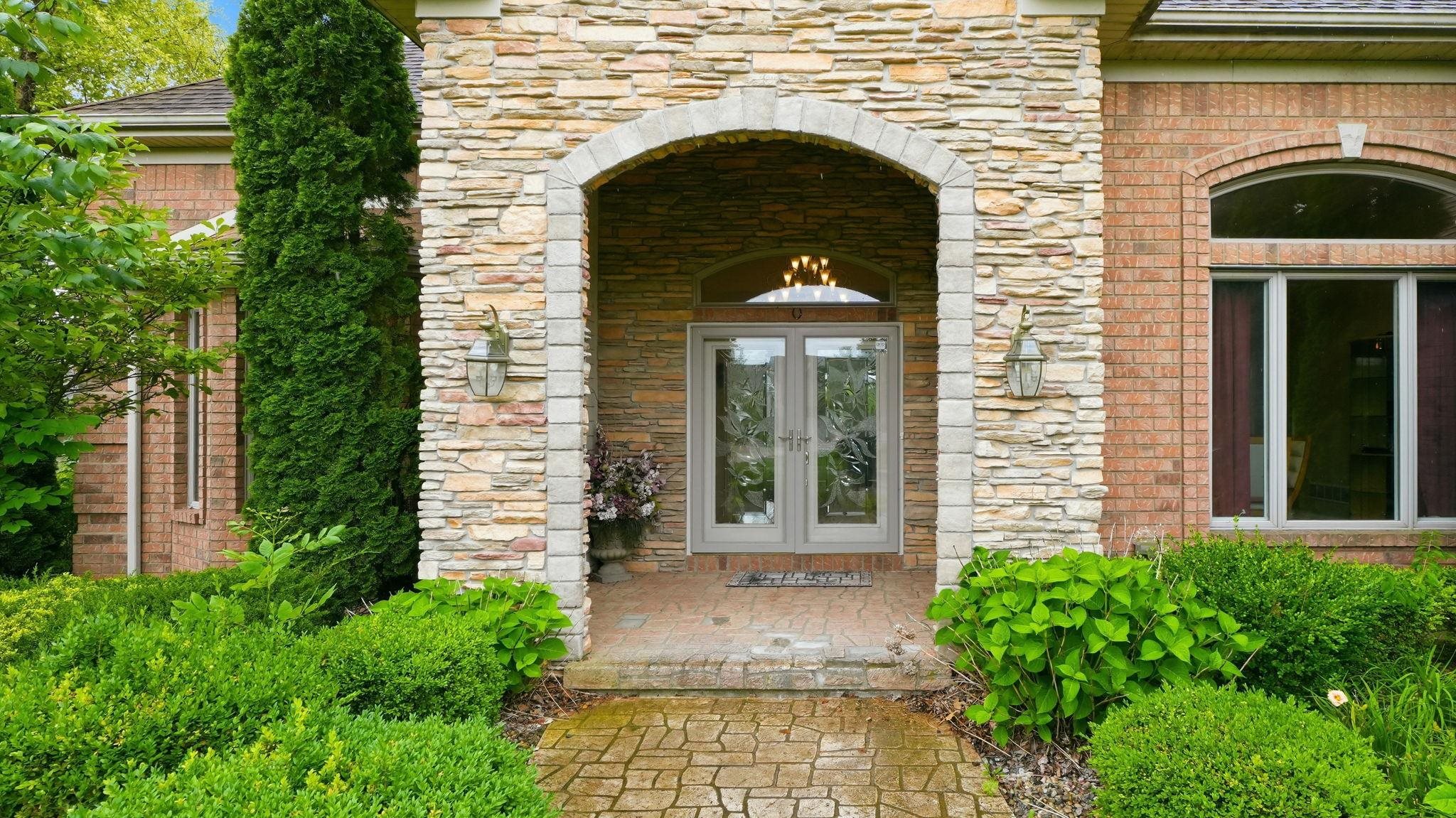 Stone and brick facade featuring an arched entryway with double glass doors, exterior sconce lighting, and a paved brick pathway framed by established green landscaping
