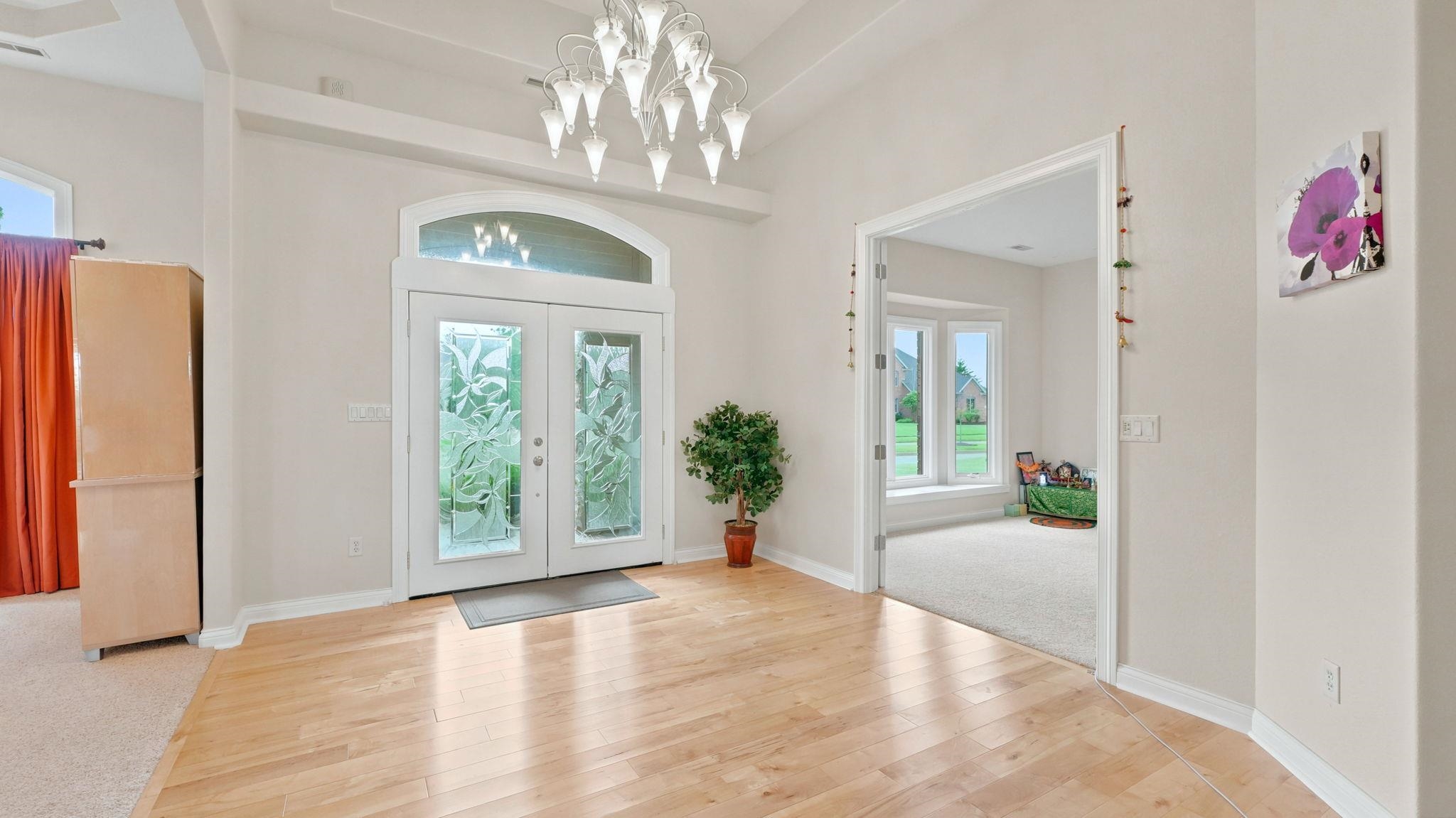 Entryway featuring light wood-finish flooring, a contemporary chandelier, double entry doors with decorative glass inserts, an arched transom window, and an open doorway to a carpeted room with bay windows