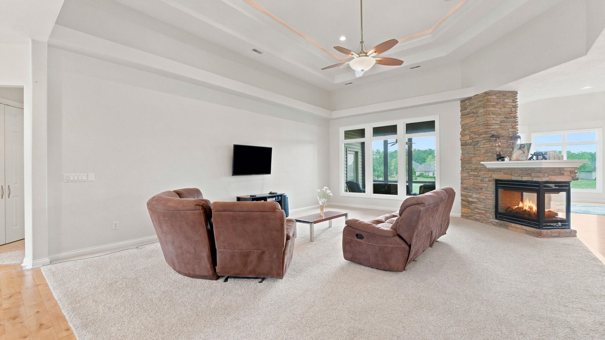 Spacious living area featuring a stone-clad fireplace with a mantel, large windows, a tray ceiling, and a ceiling fan