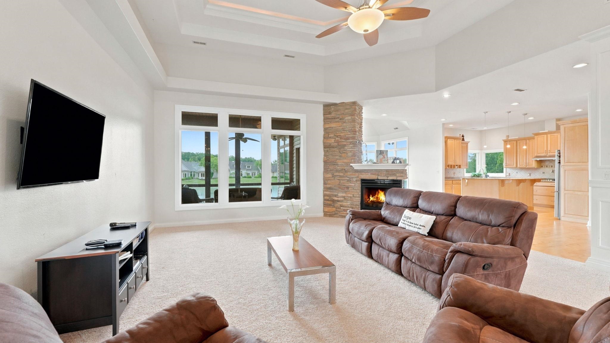 Spacious living area featuring a tray ceiling with a ceiling fan, a stone-clad fireplace with a mantel, and large windows overlooking a body of water
