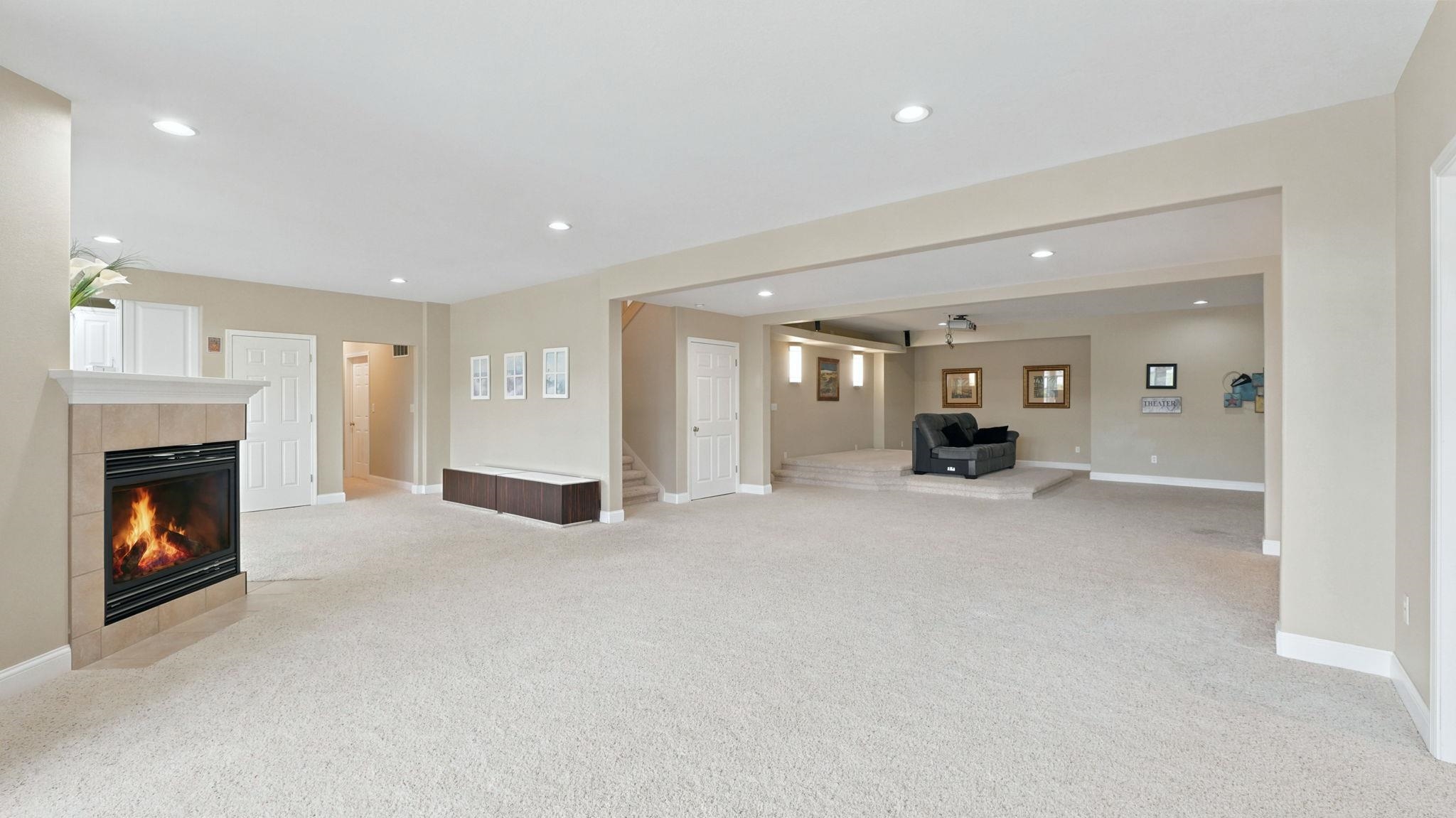 Spacious recreational room featuring a fireplace with a light-toned tile surround and mantle, recessed lighting, neutral carpeting, and a raised platform area
