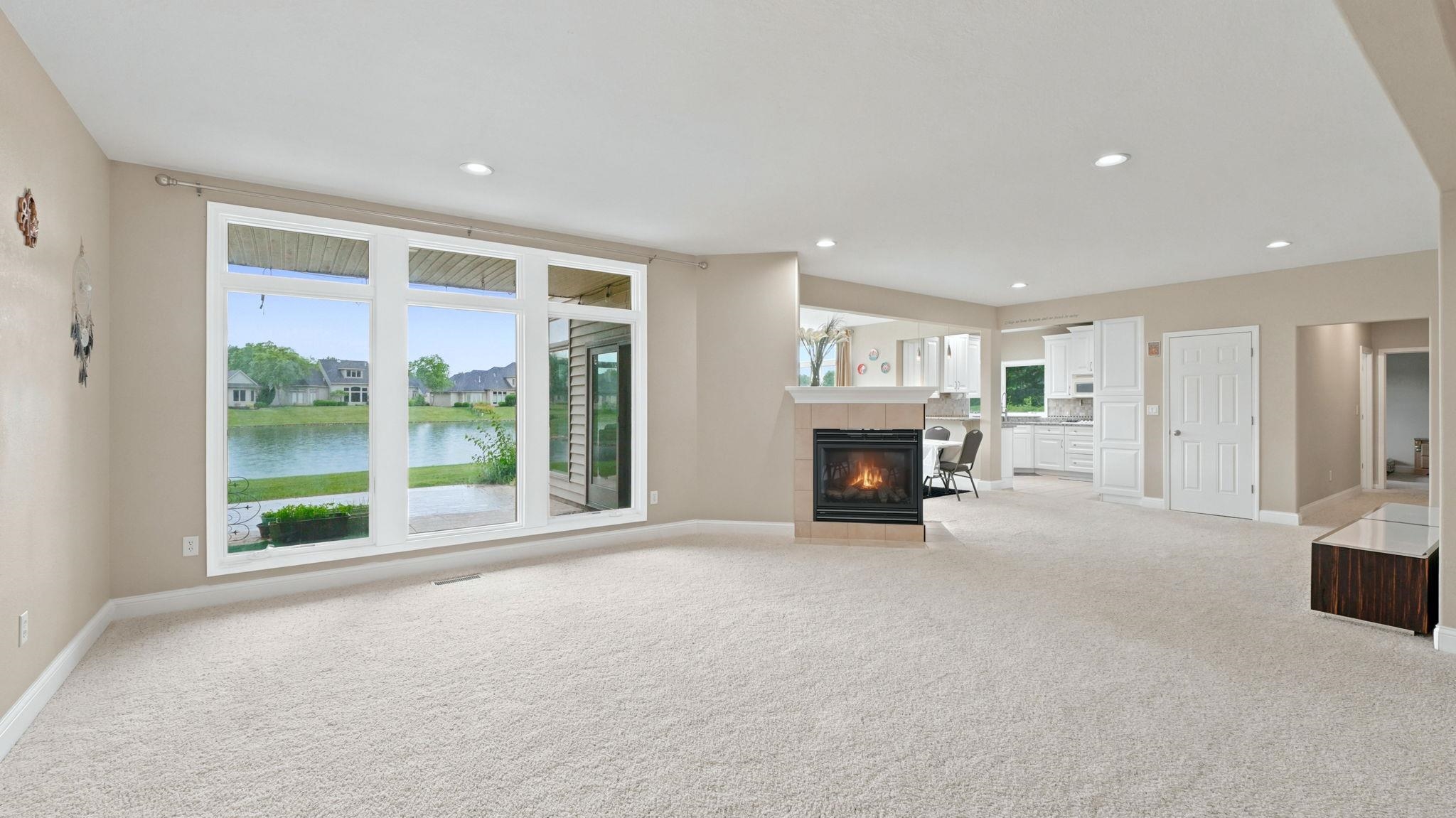 Open-concept living space featuring a corner fireplace with a tiled surround, recessed lighting, and expansive windows offering a water feature backdrop