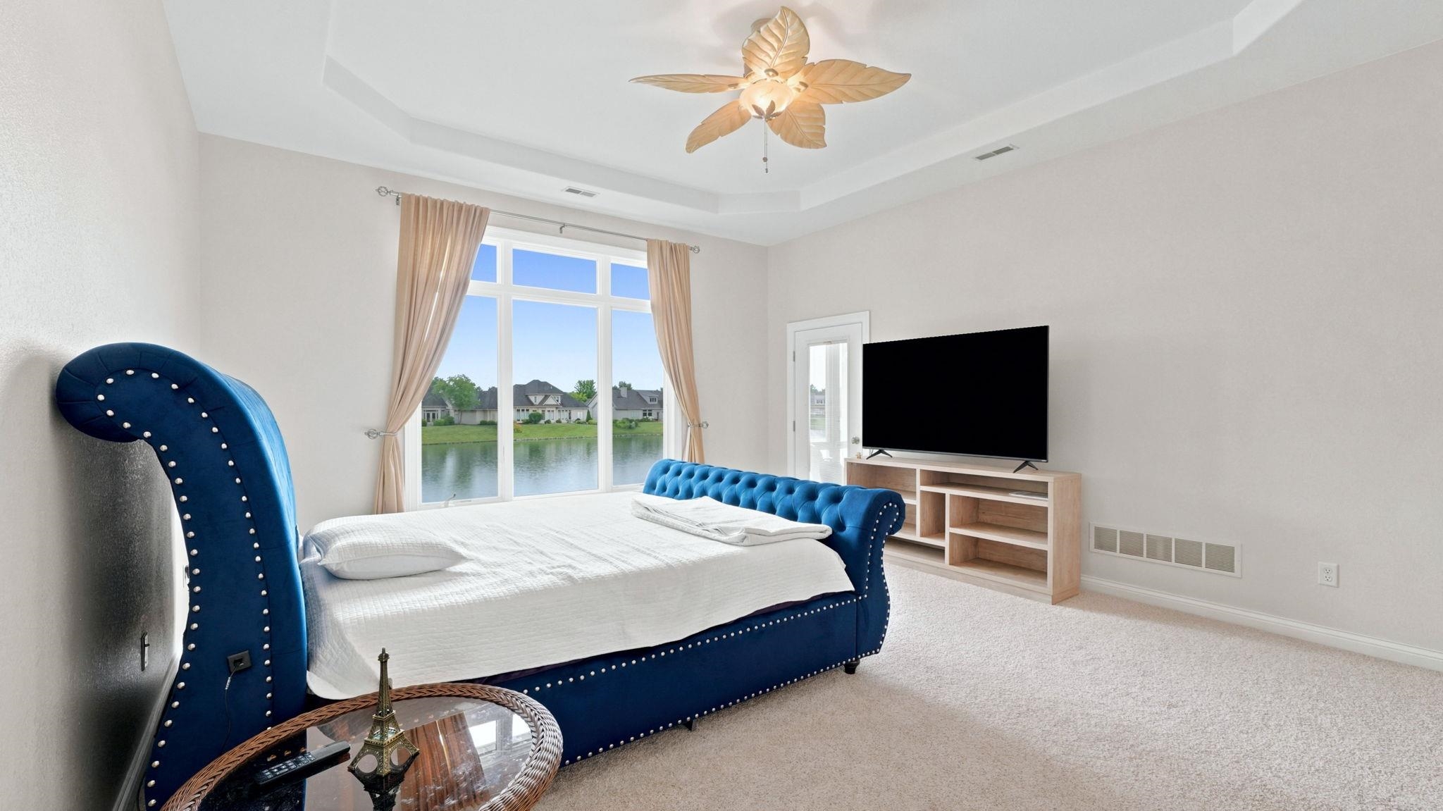 Spacious carpeted room featuring a double tray ceiling, a tropical-inspired ceiling fan, and a large window overlooking a body of water