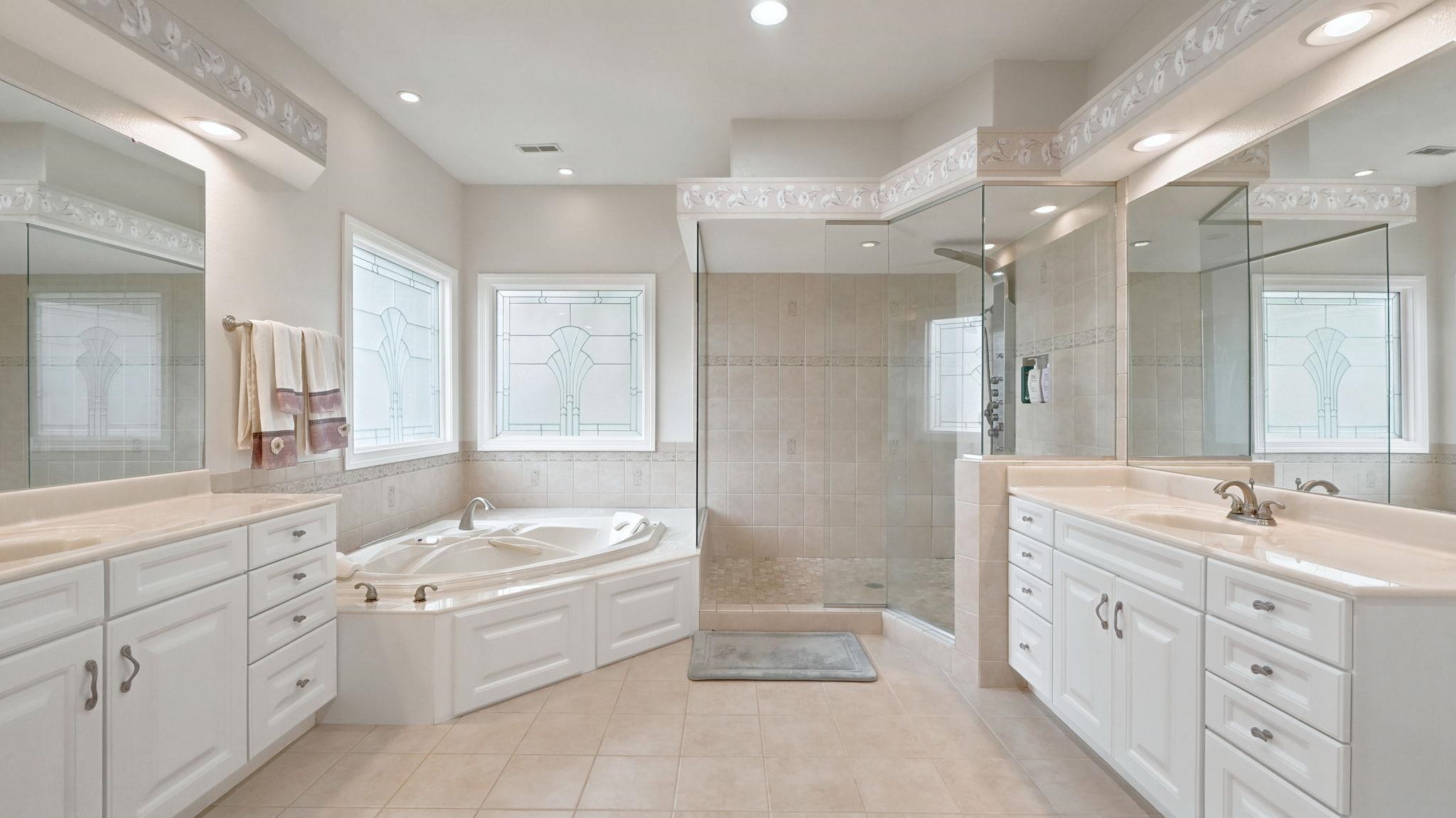 Spacious bathroom featuring a corner soaking tub, dual vanities with integrated sinks, a glass-enclosed shower, and decorative windows