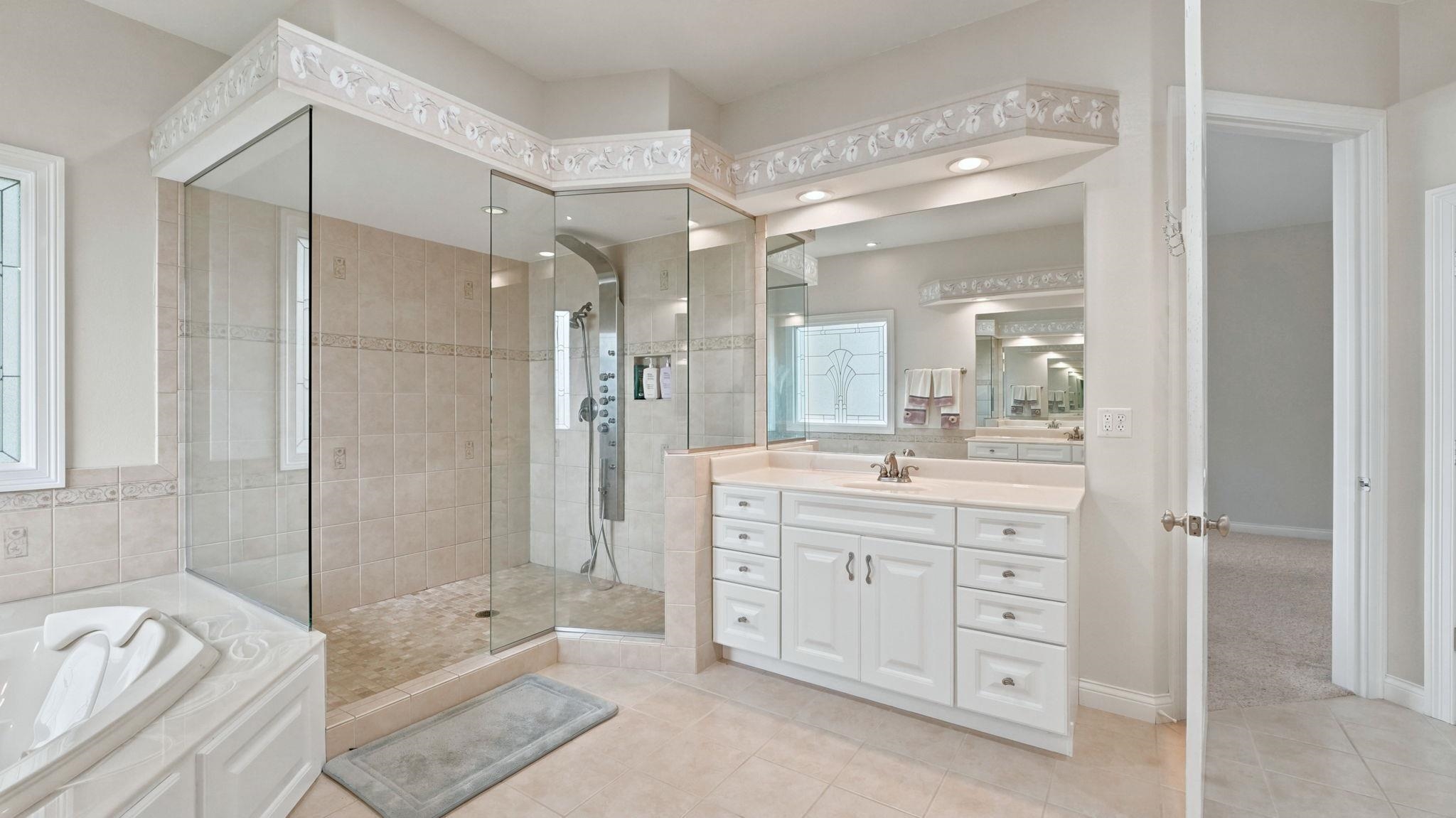 Spacious bathroom featuring a glass-enclosed shower with multiple showerheads, a built-in tub with tile surround, a large vanity with ample storage, a wide mirror, and recessed lighting