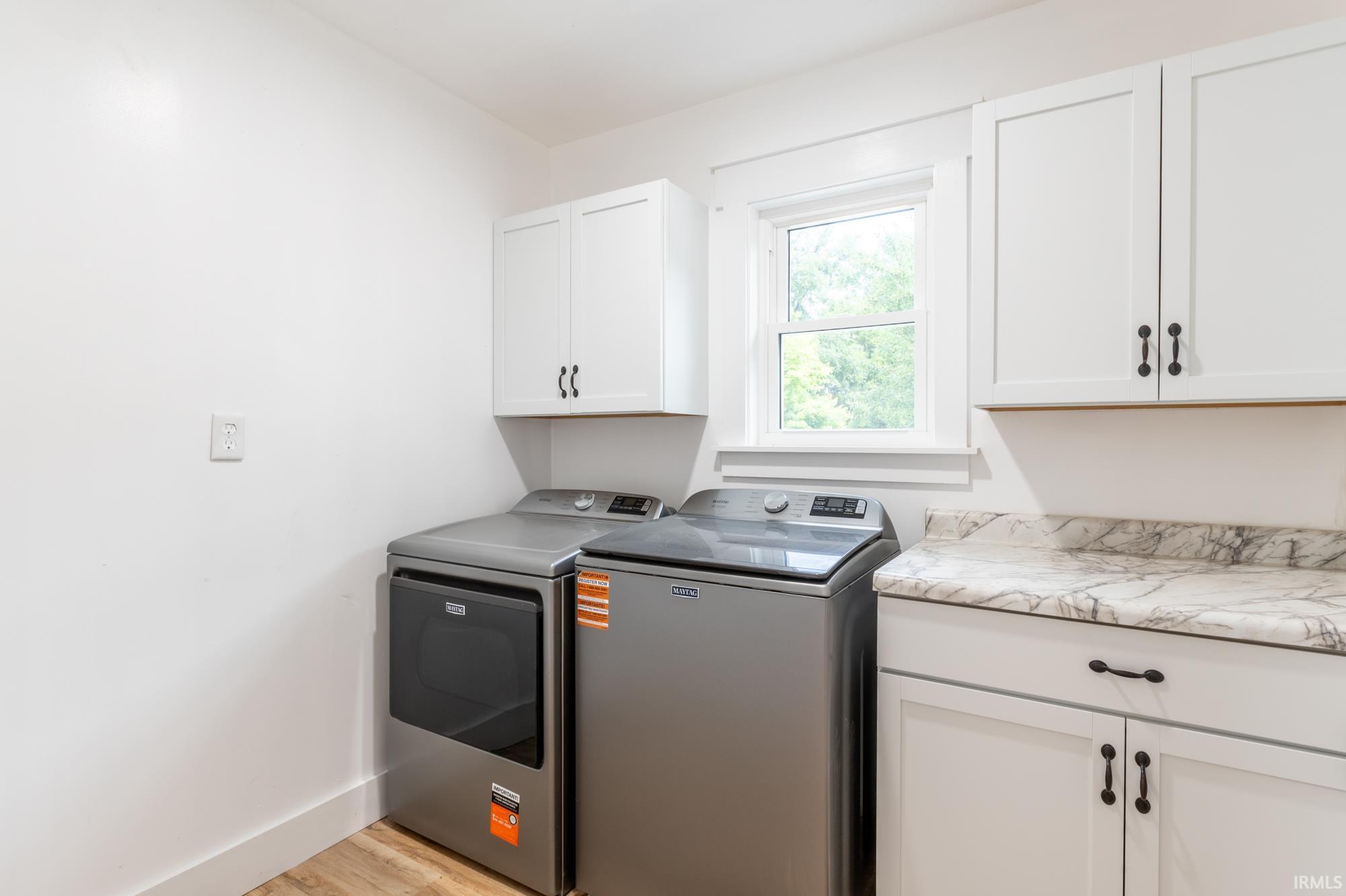 Dedicated laundry area featuring a window, wood-finish flooring, white shaker-style cabinetry with dark hardware, and a light-toned countertop