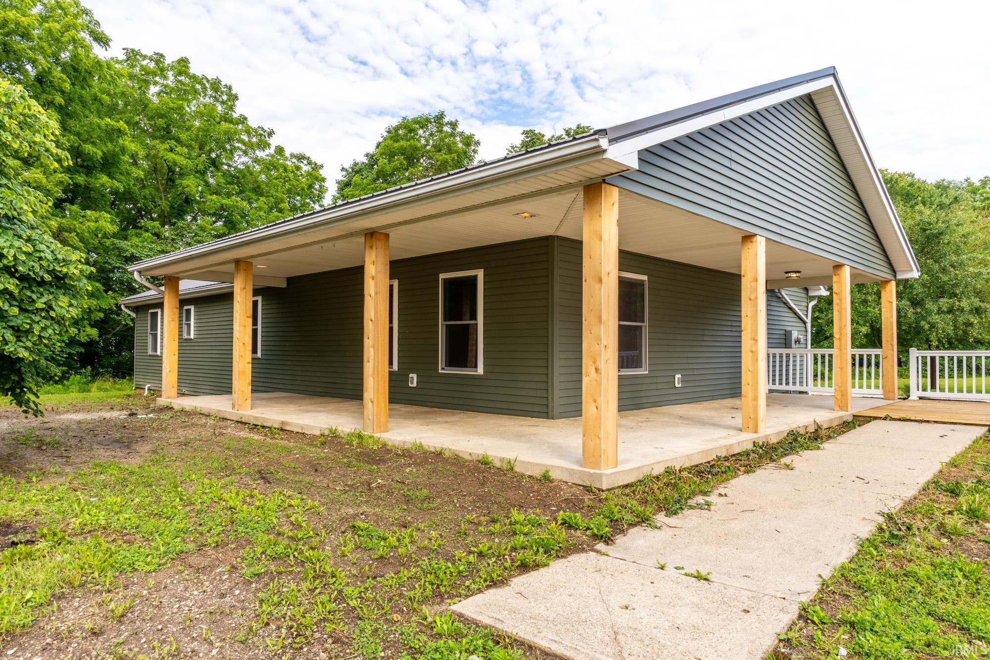 House exterior featuring dark green horizontal siding, a covered concrete porch, natural wood support columns, a gray metal roof, and white-framed windows