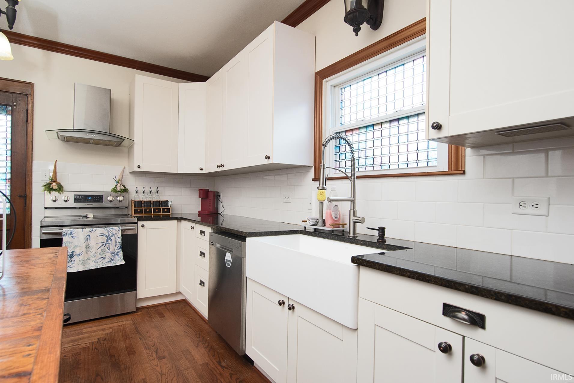 Kitchen featuring white shaker-style cabinetry, dark countertops, a farmhouse sink, stainless steel appliances, and wood-finish flooring
