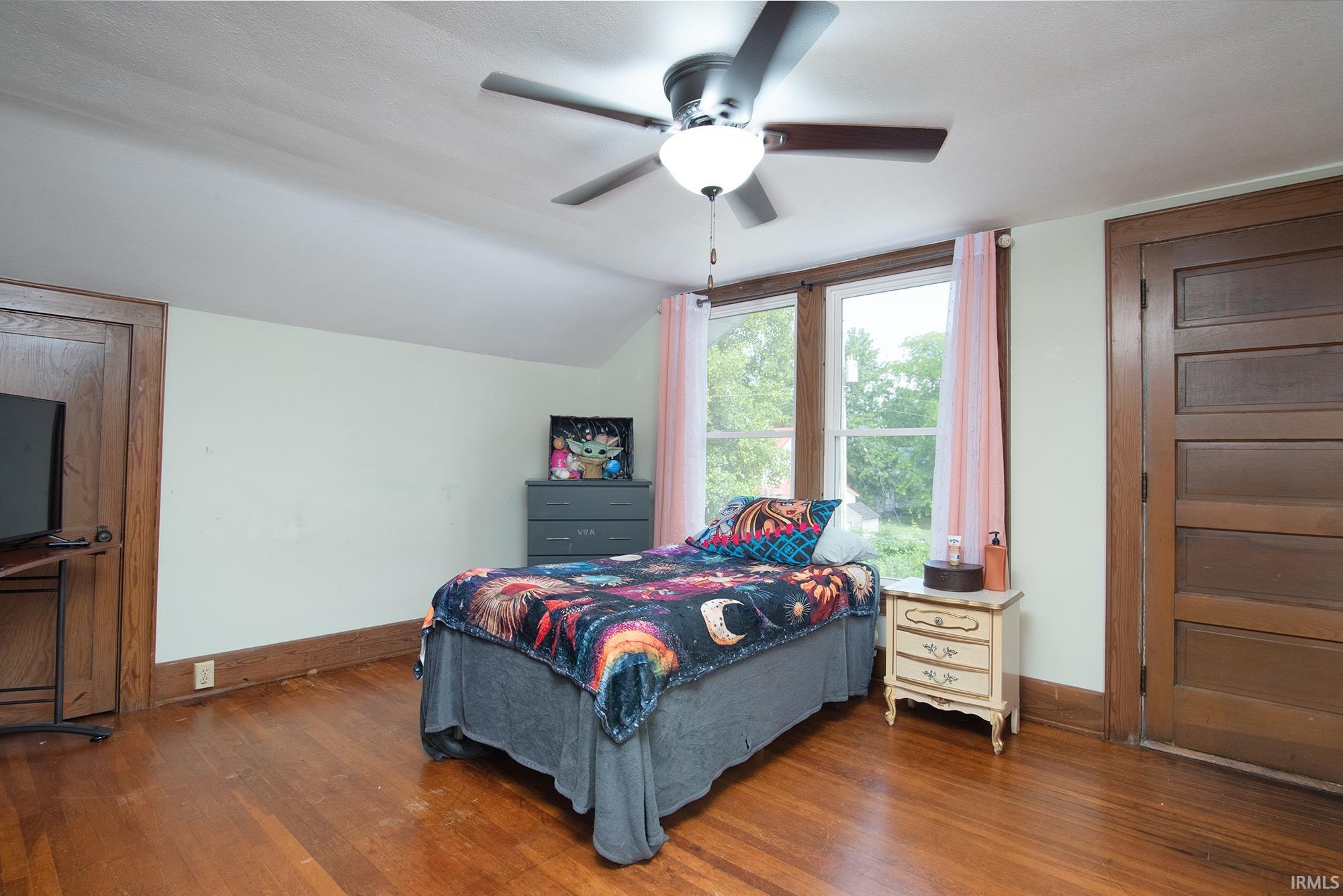 Bedroom featuring rich wood-finish flooring, a ceiling fan with integrated lighting, and multiple windows with wood trim