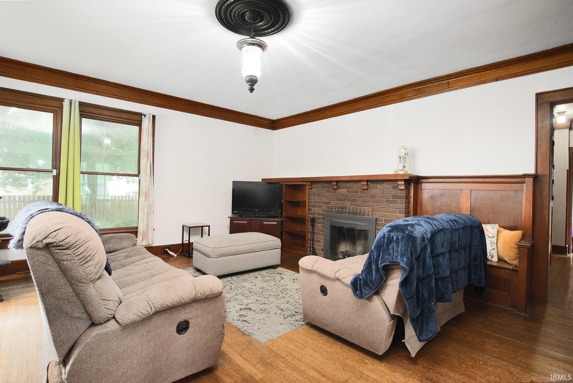 Living area featuring a brick fireplace with a wood mantle, built-in shelving, hardwood flooring, and decorative crown molding