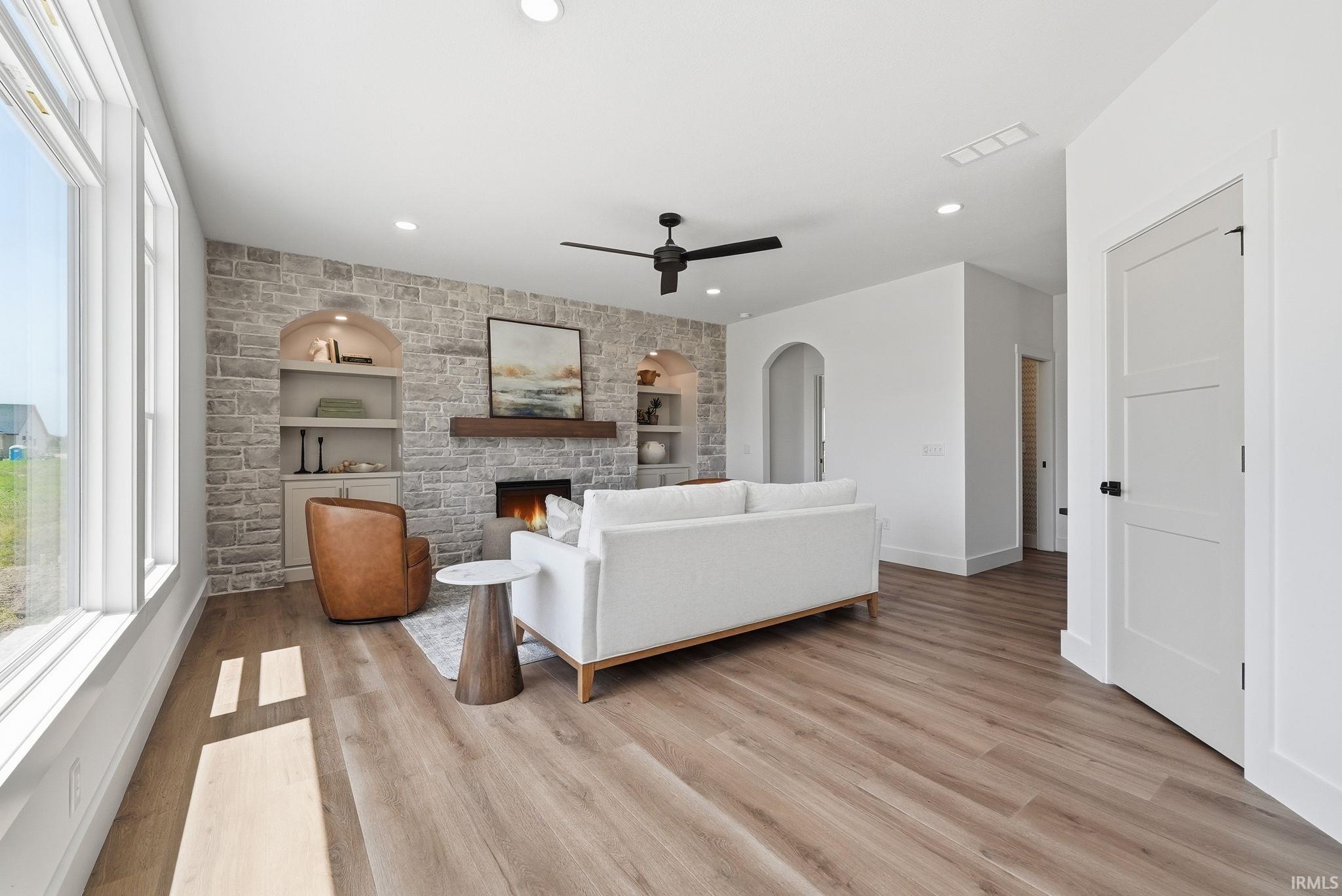 Spacious living area featuring stone-finish fireplace with wood mantel, built-in shelving, wood-finish flooring, and ample natural light from large windows