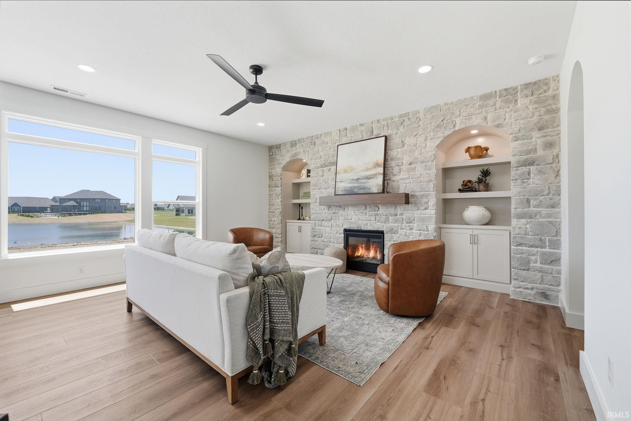 Spacious living area featuring large picture windows, wood-finish flooring, a stone accent wall, an electric fireplace with a wood mantel, and arched built-in shelving with lower cabinetry