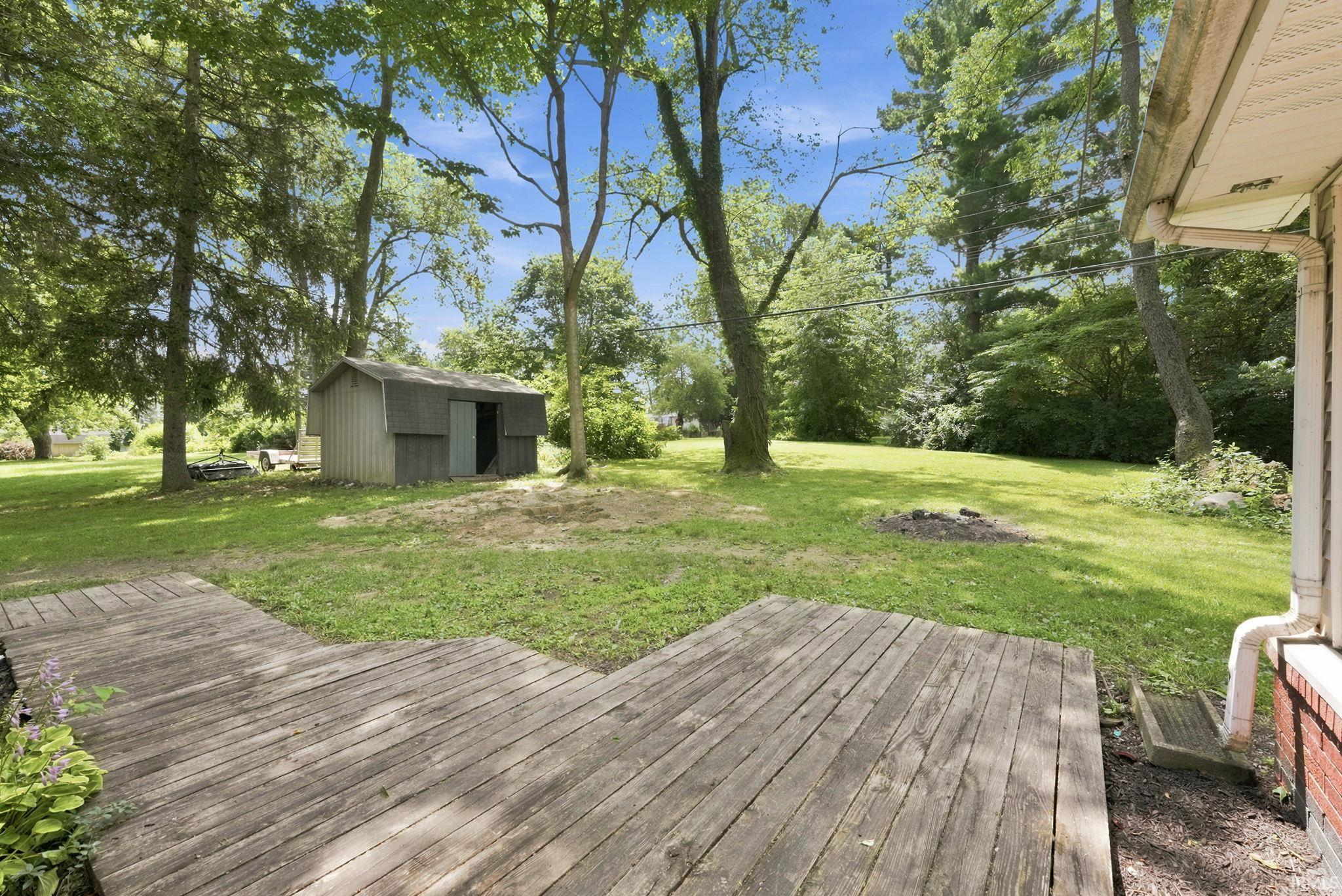 Expansive backyard featuring a large wooden deck, mature trees, a storage shed, and a fire pit