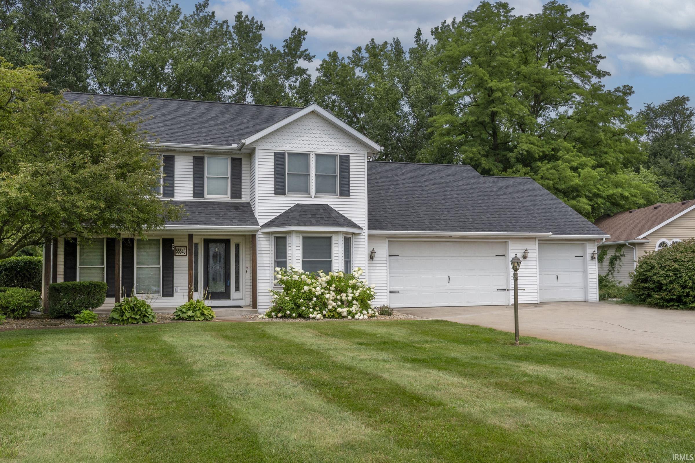 Two-story residence featuring white siding and dark shutters, a three-car garage, established front landscaping, a bay window, and a spacious front lawn