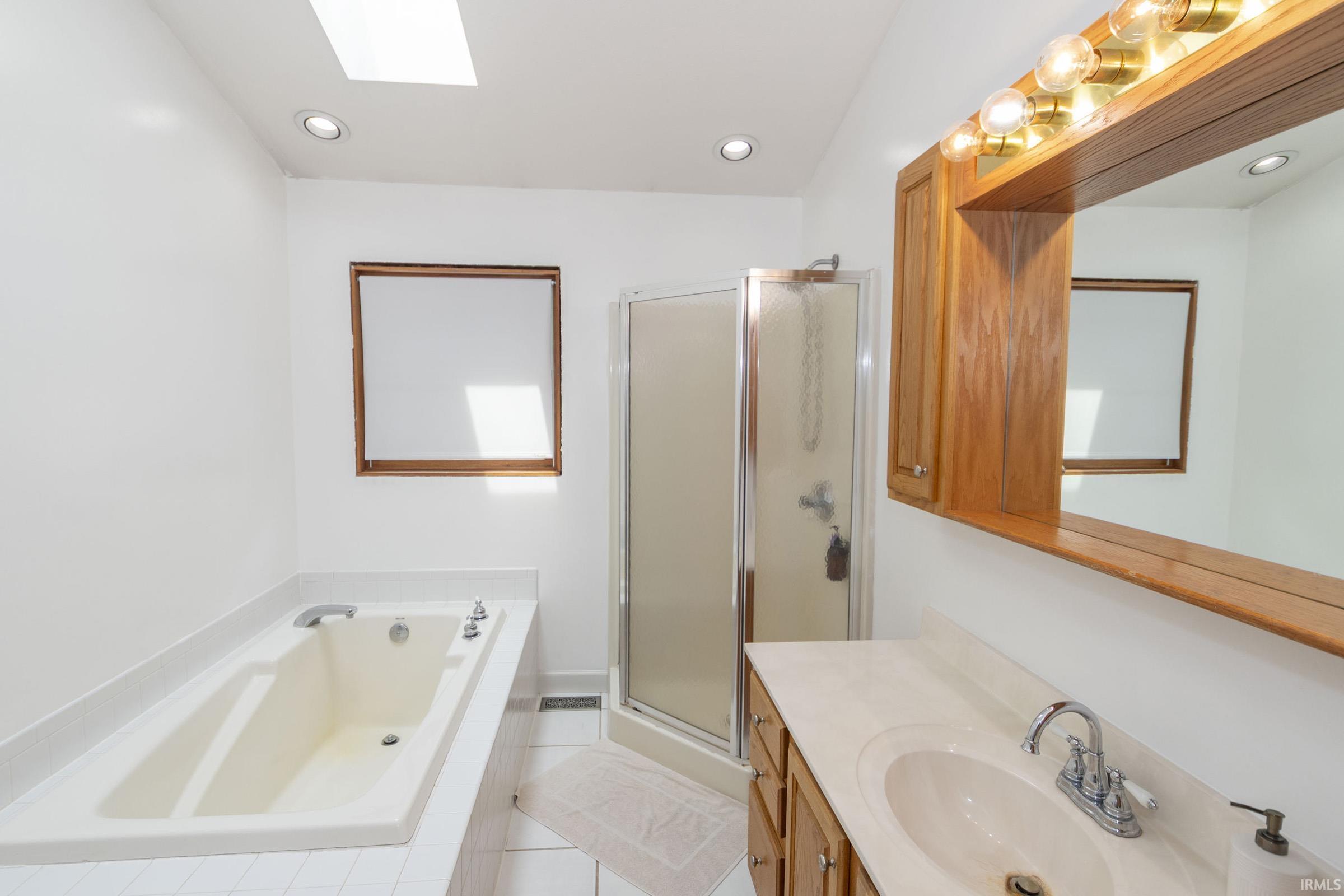 Bathroom featuring a drop-in soaking tub, an enclosed shower, a wood-finish vanity with an integrated sink, a large wall-mounted mirror, and a ceiling skylight