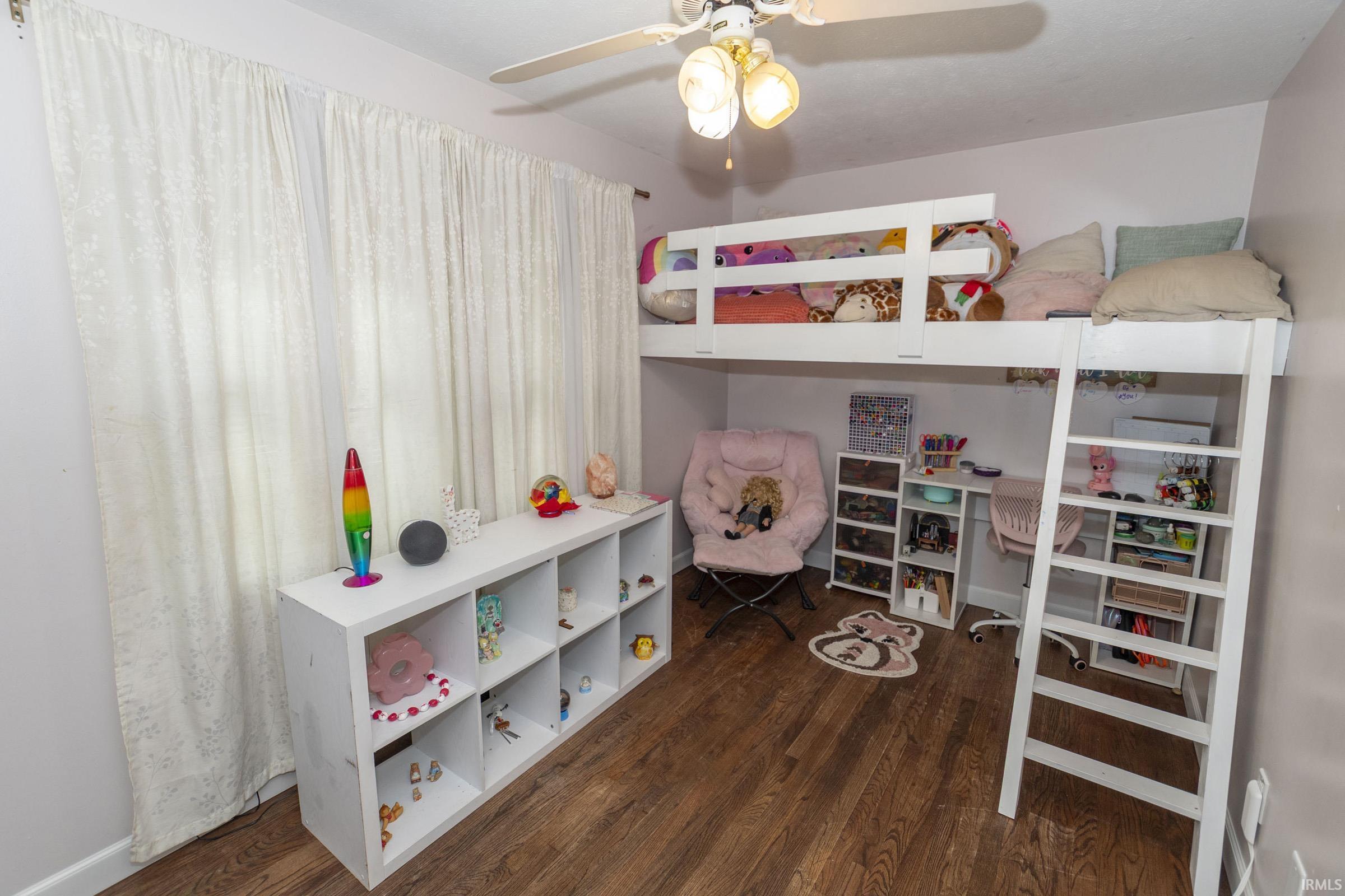 Room featuring wood-finish flooring, a white built-in loft bed, a white multi-shelf storage unit, white window treatments, and a ceiling fan with integrated lighting