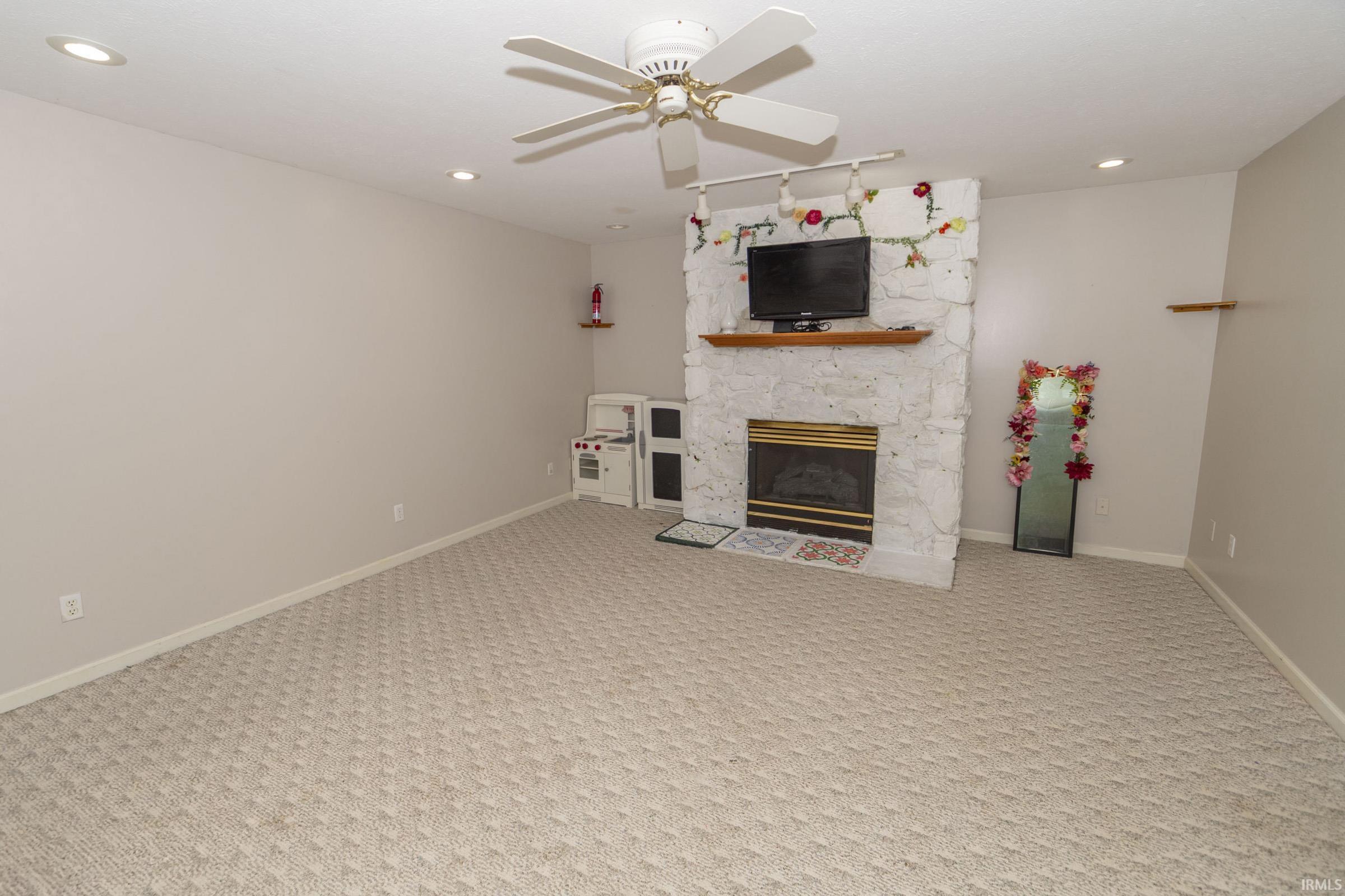 Spacious room featuring light-toned carpet flooring, a white stone fireplace with a wood mantel, and a ceiling fan with integrated lighting