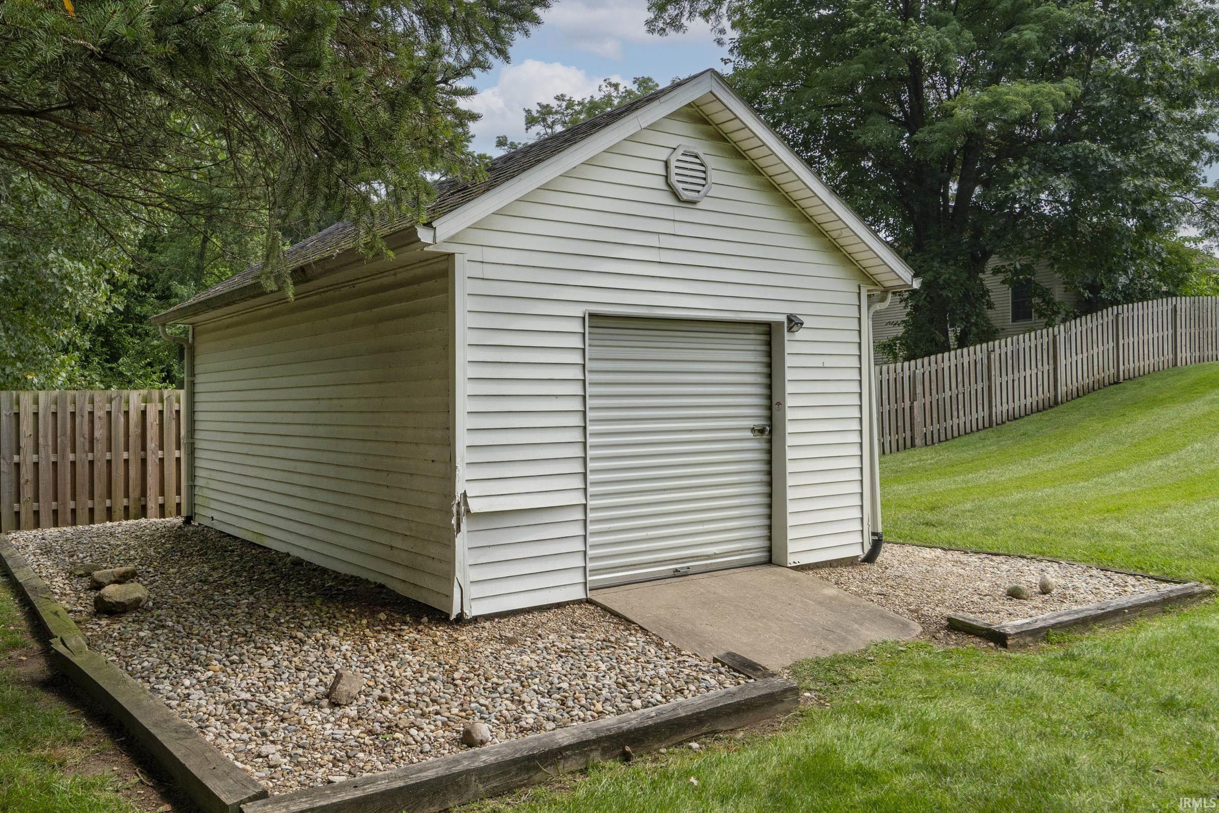 Exterior storage shed featuring white vinyl siding, an overhead garage door, a gable vent, and a concrete ramp