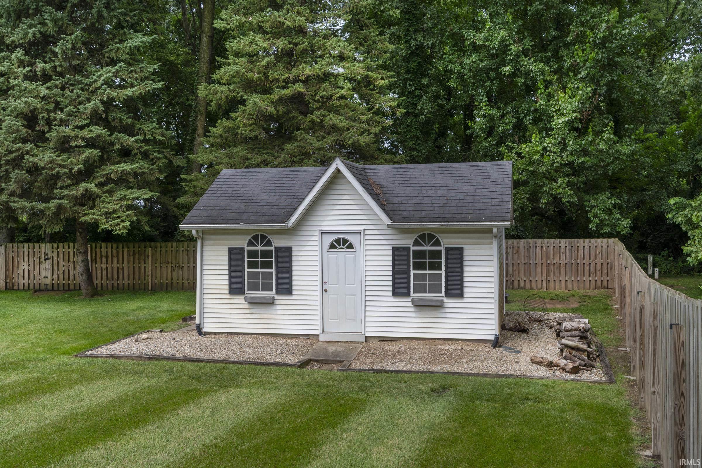 White utility structure featuring a gabled roof, arched windows with dark shutters, a panel door, and a gravel perimeter