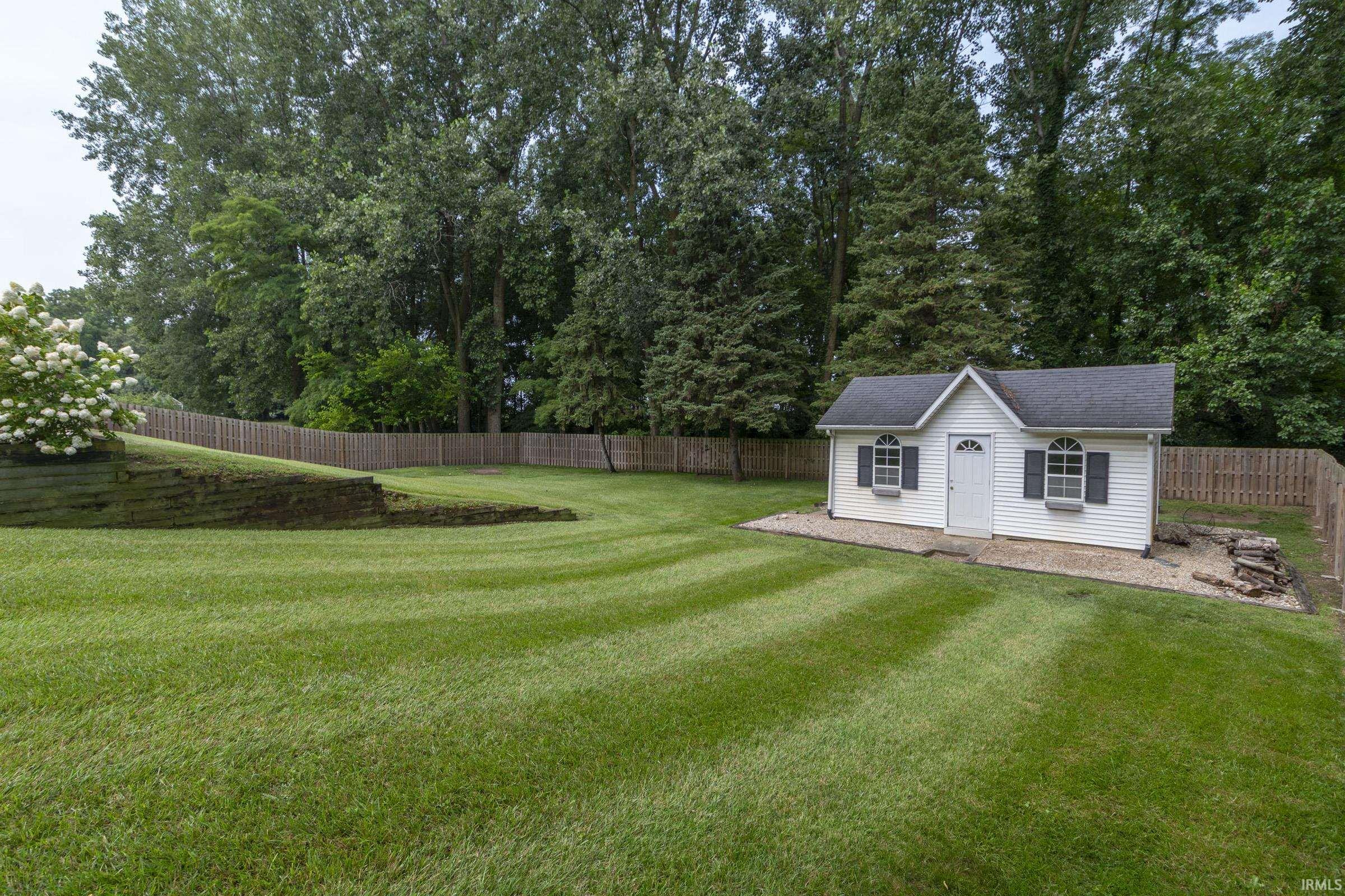 Expansive lawn area featuring a storage shed with arched windows and window boxes, dark shutters, and a wood privacy fence