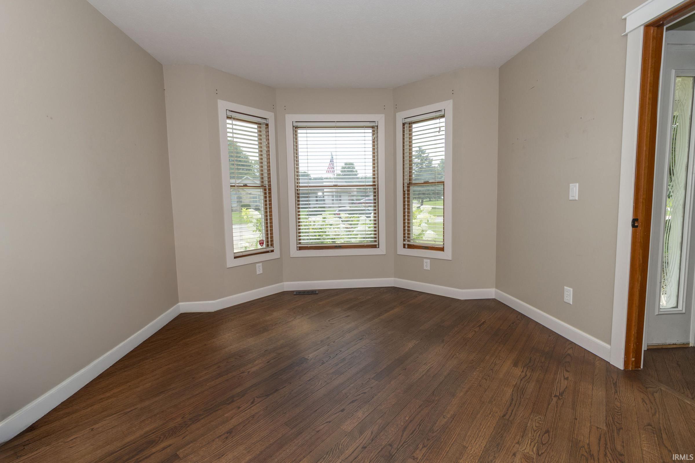 Bay window with three individual windows, wood-finish flooring, light-toned wall paint, white baseboards, and a wood-trimmed doorway