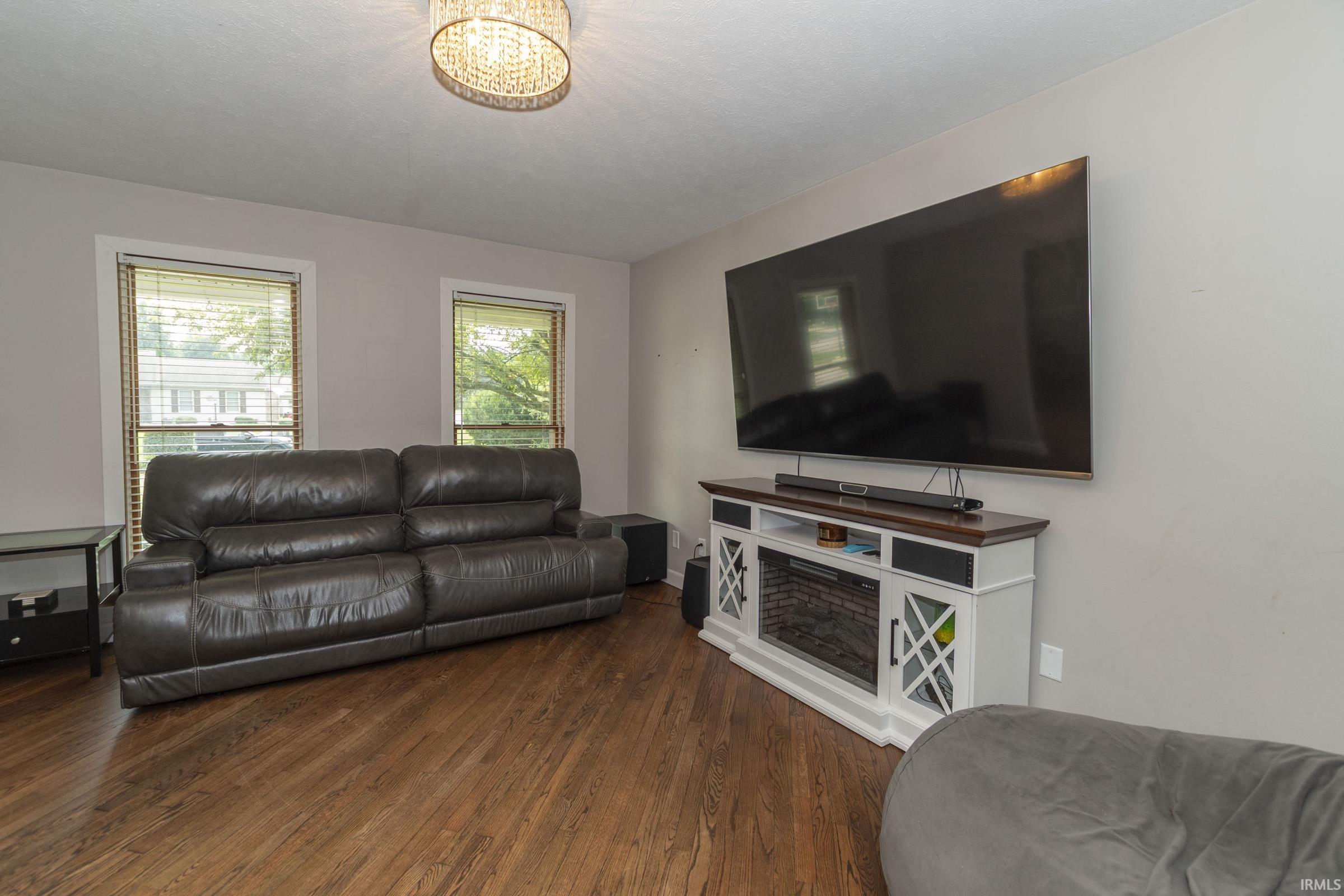 Spacious living area featuring wood-finish flooring, two windows with blinds, a decorative ceiling light fixture, a built-in media console, and light-toned walls