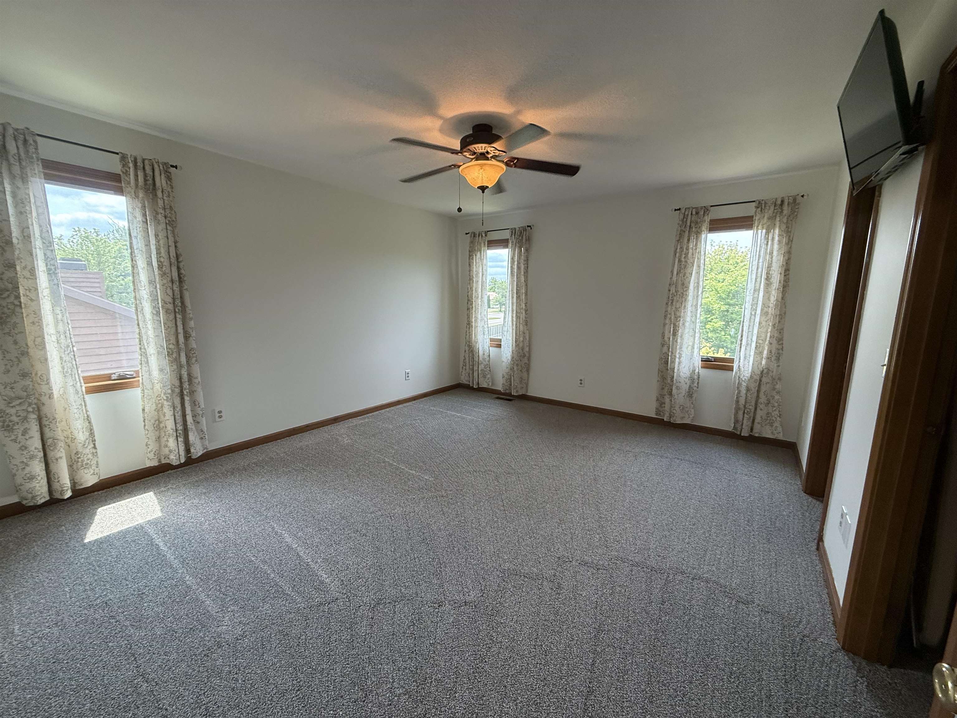 Spacious room featuring neutral carpeting, a ceiling fan with integrated lighting, and multiple windows with wood trim