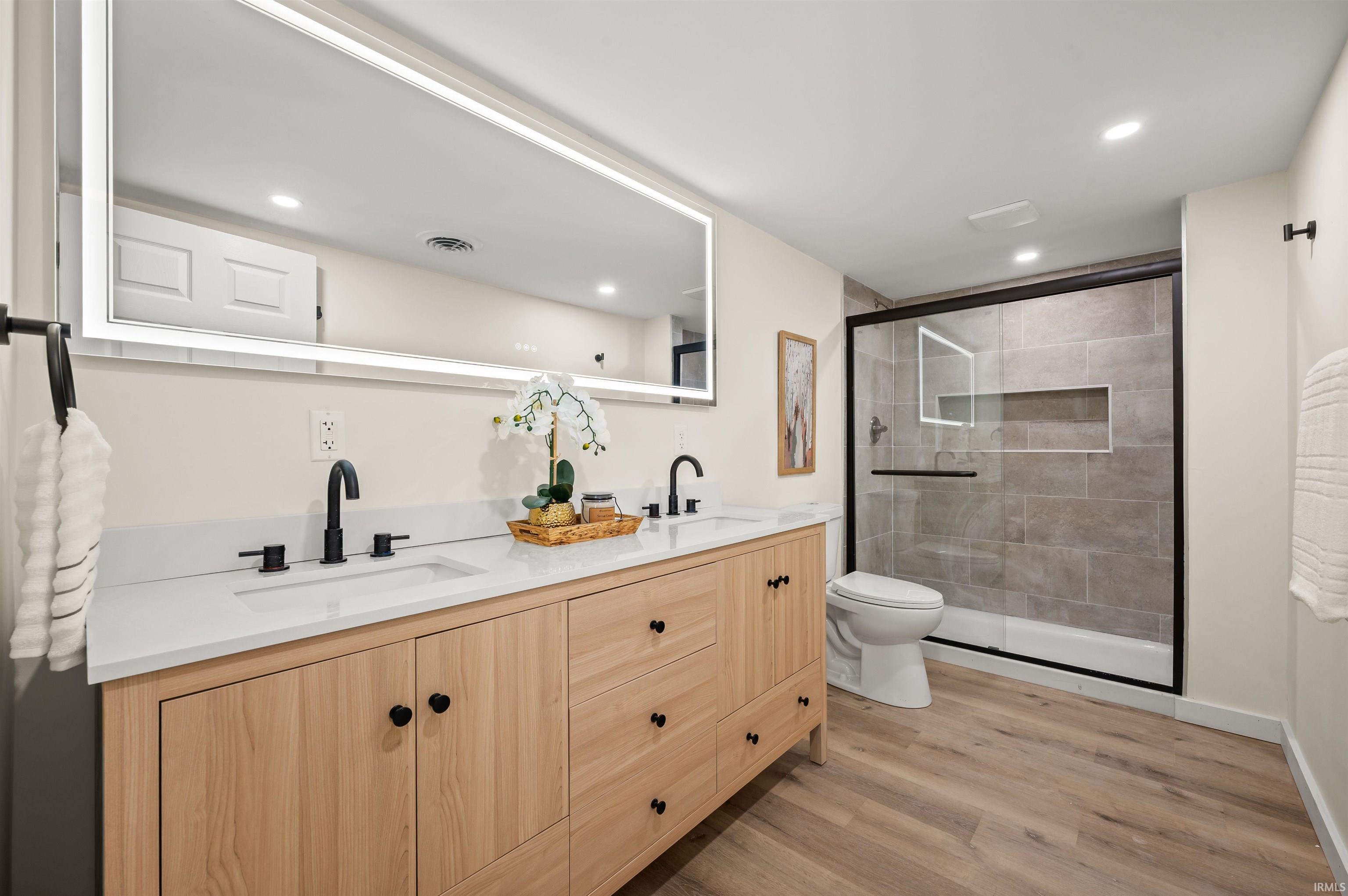 Light wood-finish double vanity with matte black fixtures, quartz countertops, and an integrated illuminated mirror