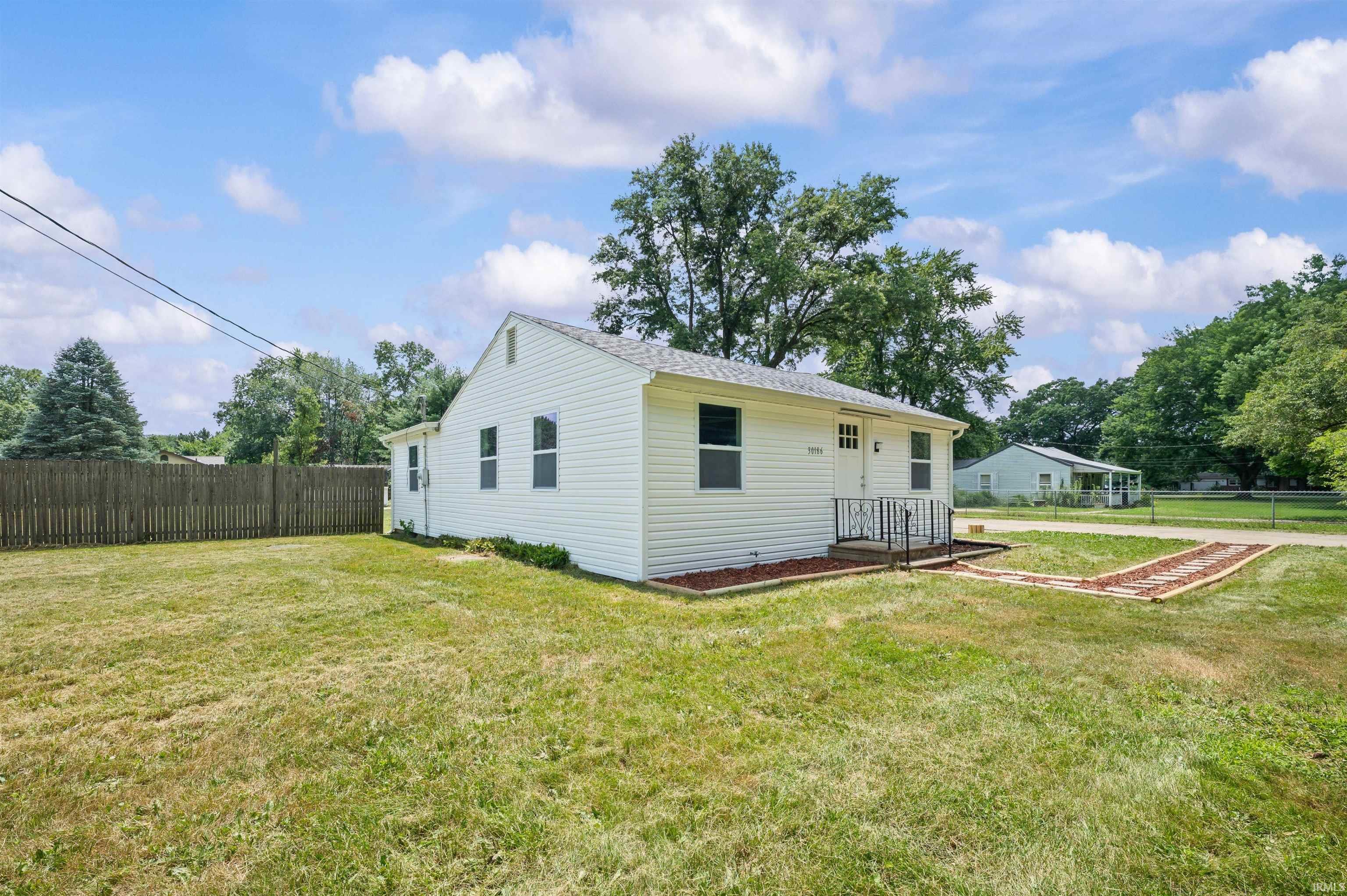 White siding exterior with a pitched roof, front entry porch with railing, wood fence, and expansive lawn area