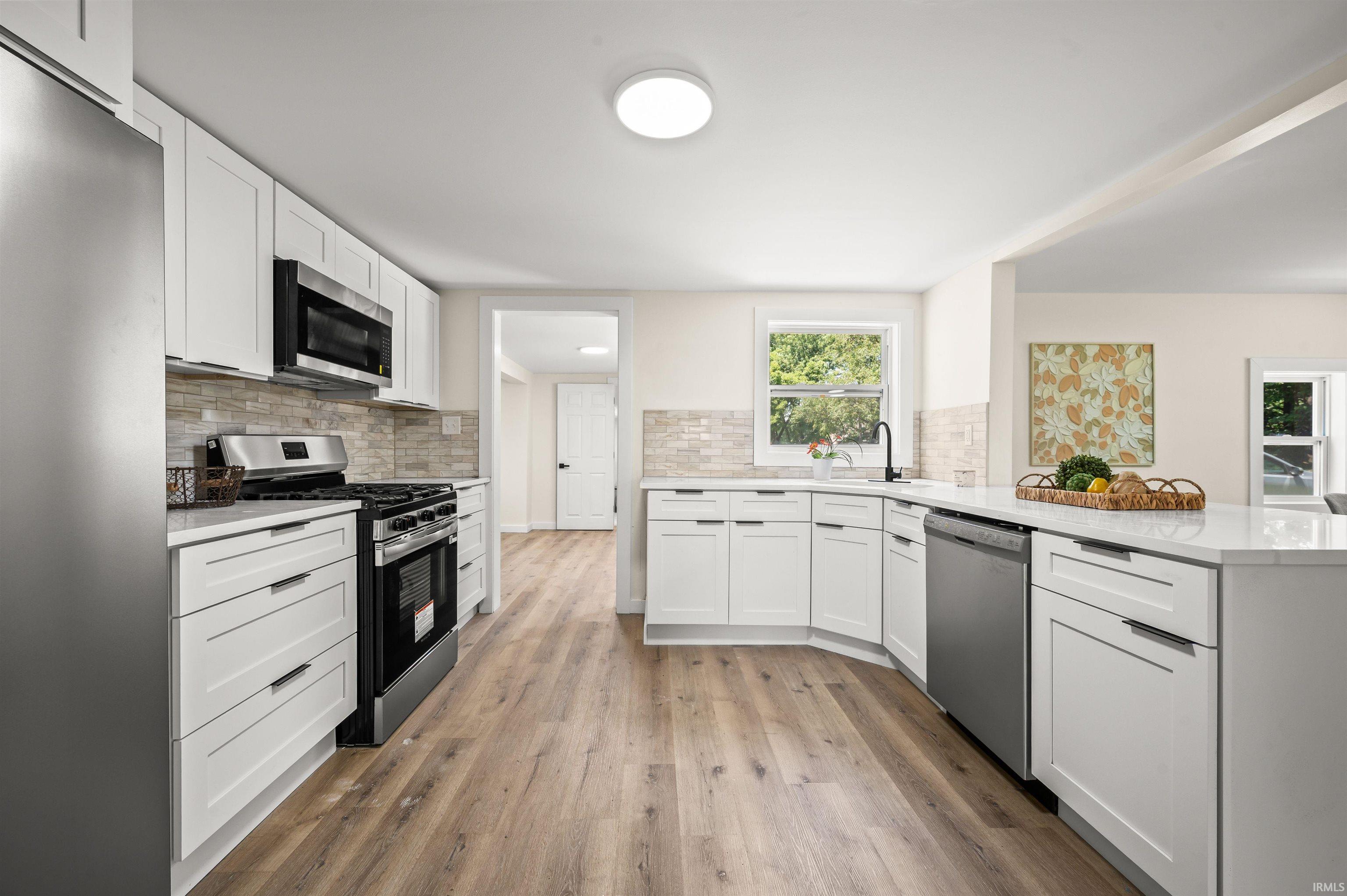 Modern kitchen featuring white shaker cabinetry, stainless steel appliances, light-toned wood-finish flooring, a stone-look backsplash, and white countertops