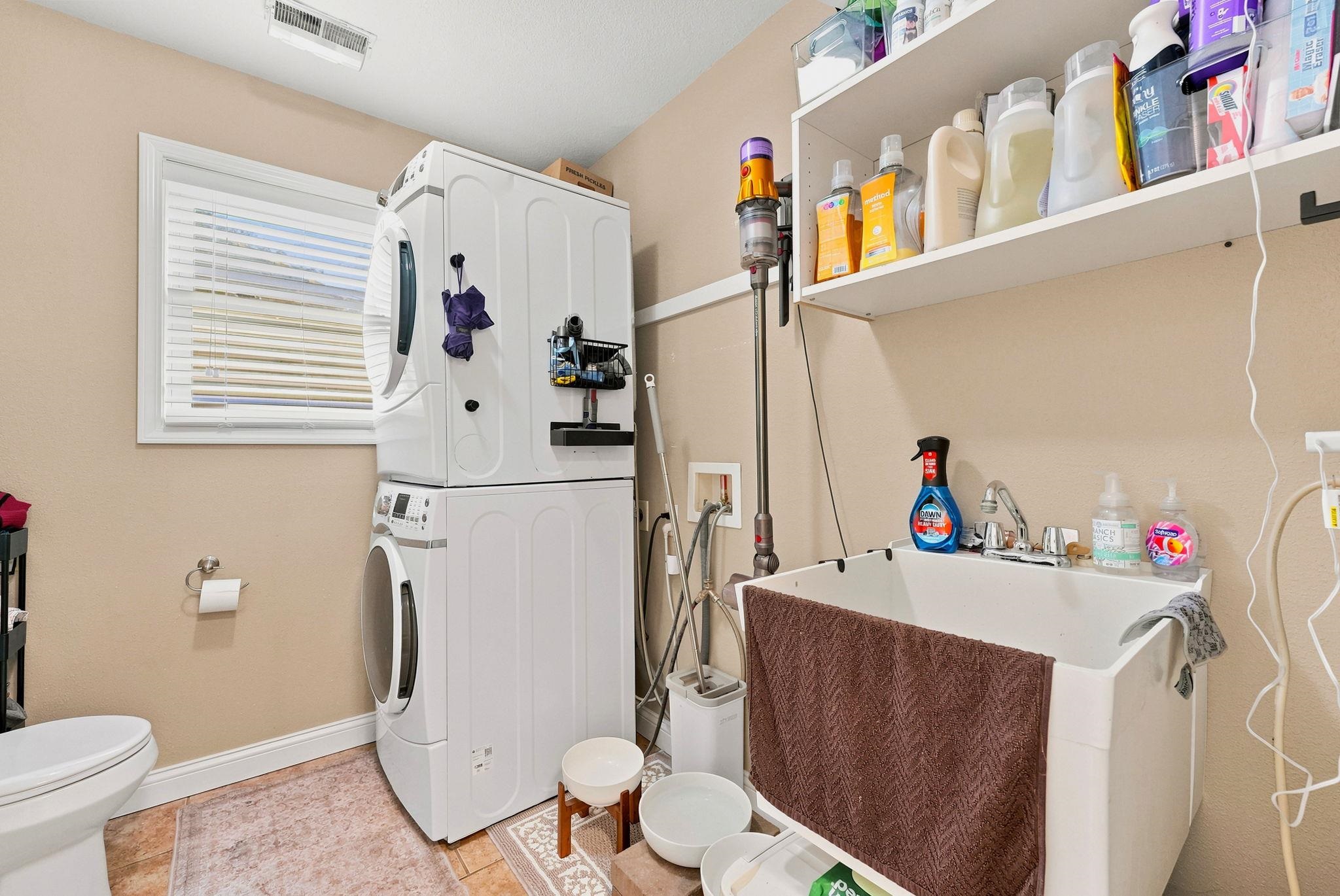 Dedicated laundry area featuring a stacked washer and dryer, a utility sink with dual faucets, and a window with horizontal blinds, and half bath off Garage entry/KT