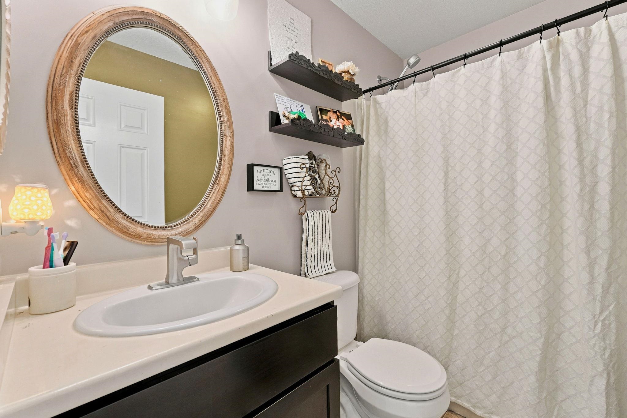 Bathroom featuring a neutral vanity countertop, undermount sink with a brushed nickel faucet, an oval mirror with a textured frame, and a shower with a black curtain rod