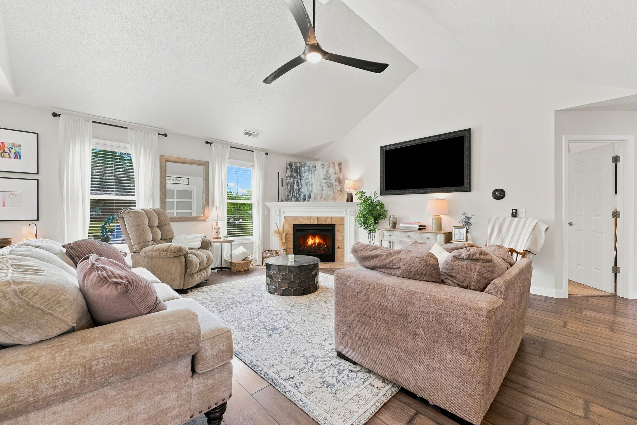 Living area featuring a vaulted ceiling with a modern ceiling fan, wood-finish flooring, a fireplace with a white mantel, and multiple windows providing natural light
